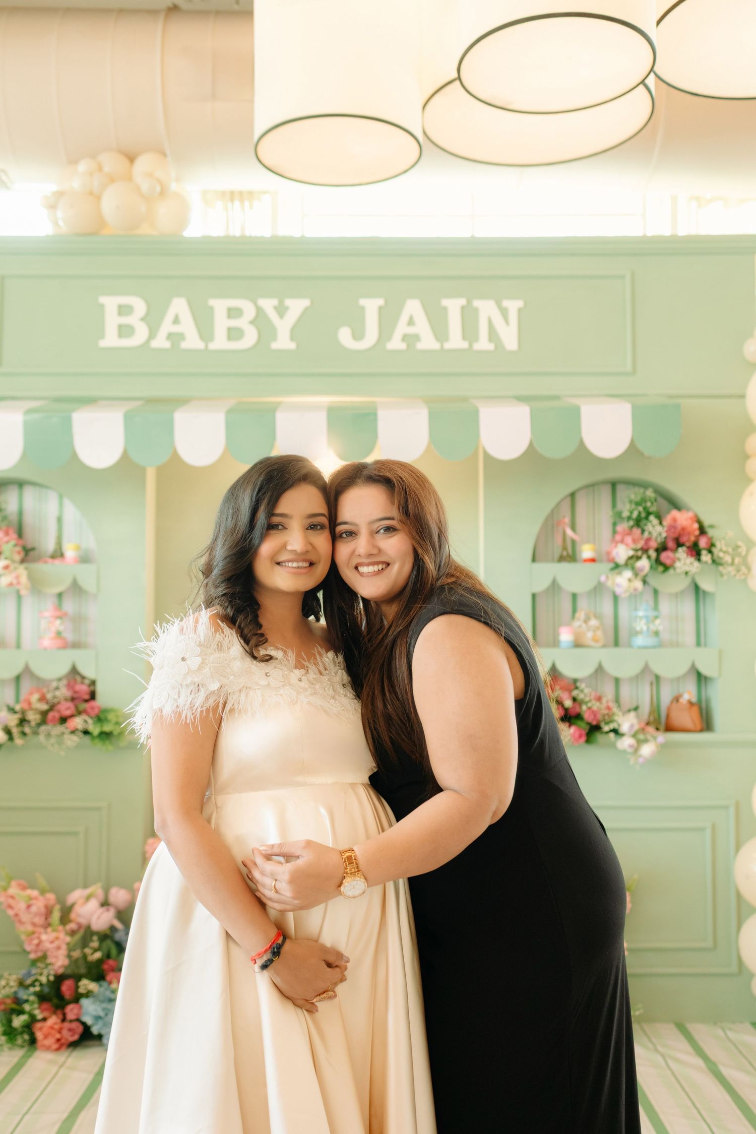 Two women pose together at a Baby Jain baby shower in front of a mint green decorative backdrop with flowers.