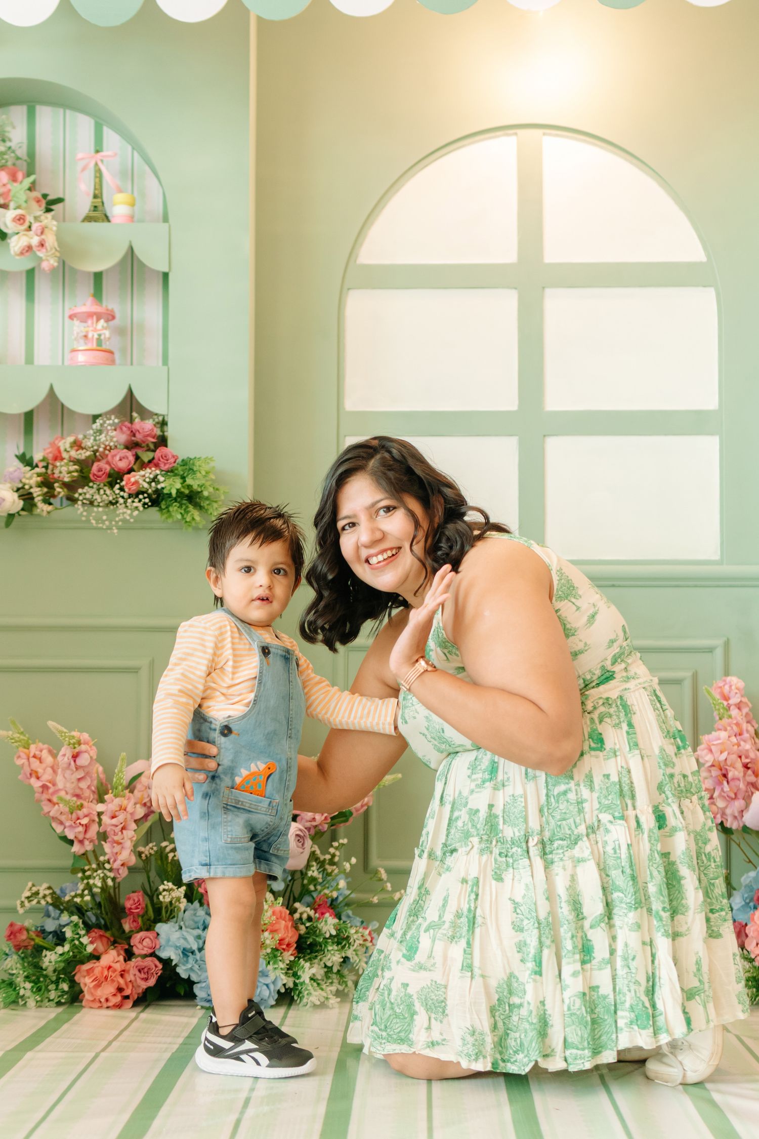 A smiling woman in a floral dress poses with a toddler boy in denim overalls in a pastel green studio setting.