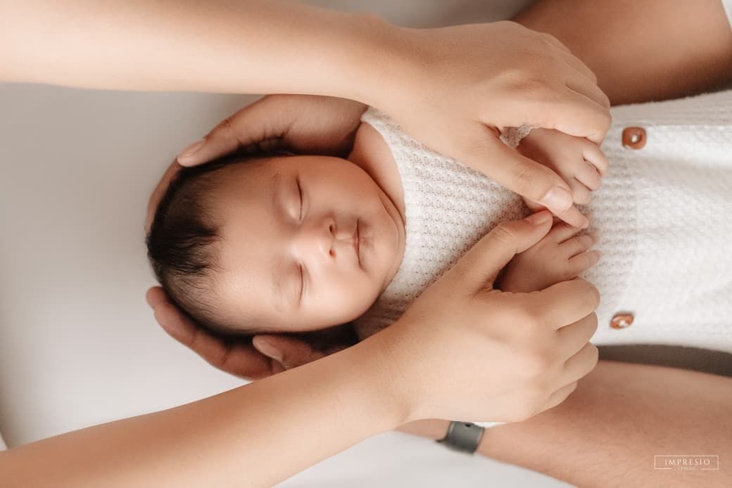 Newborn baby sleeping peacefully in white knit outfit, cradled gently by parents hands on soft surface.