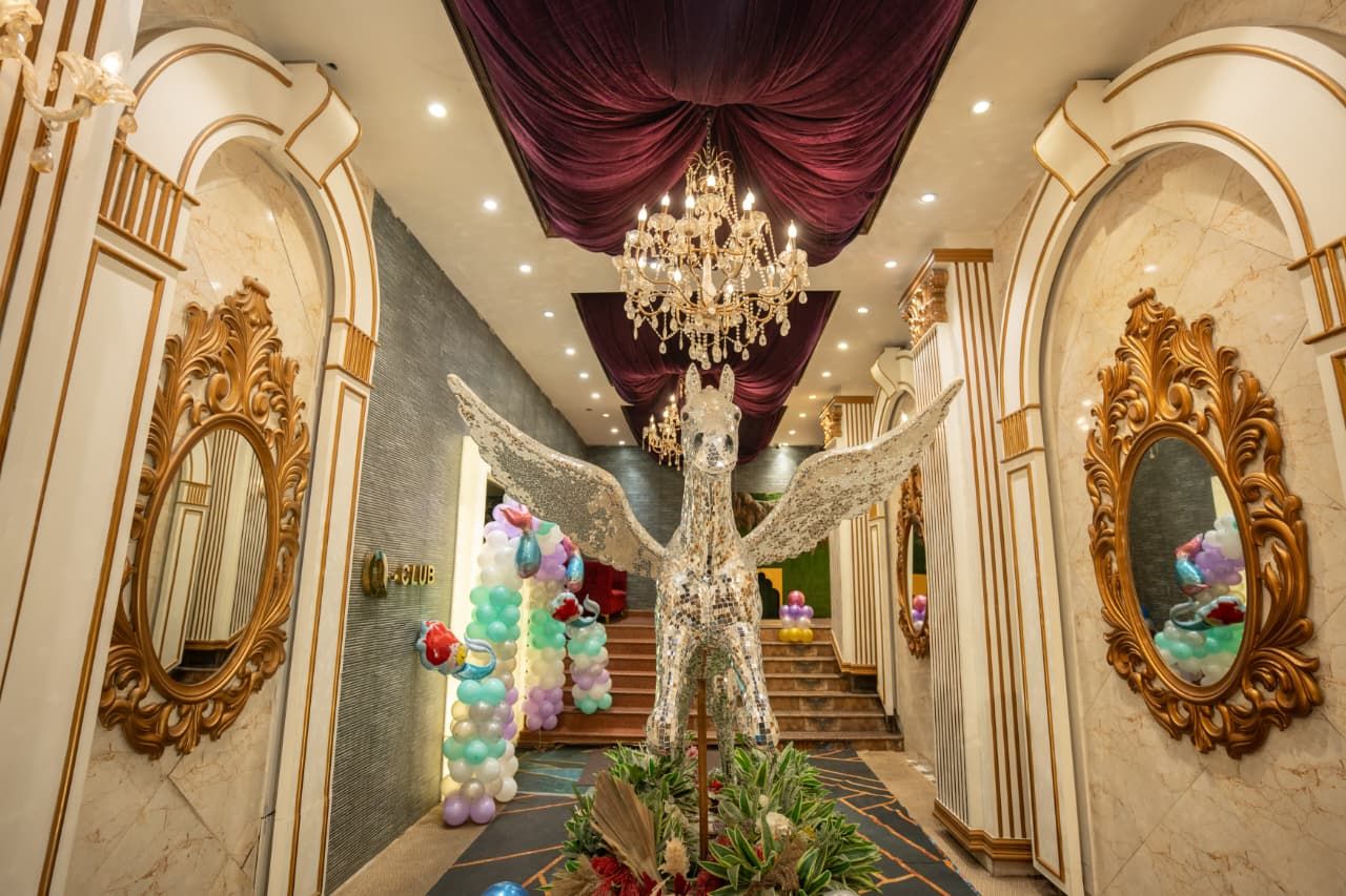 Opulent hallway with crystal chandelier, gold-framed mirrors, purple drapes, and an angel statue surrounded by colorful balloons.