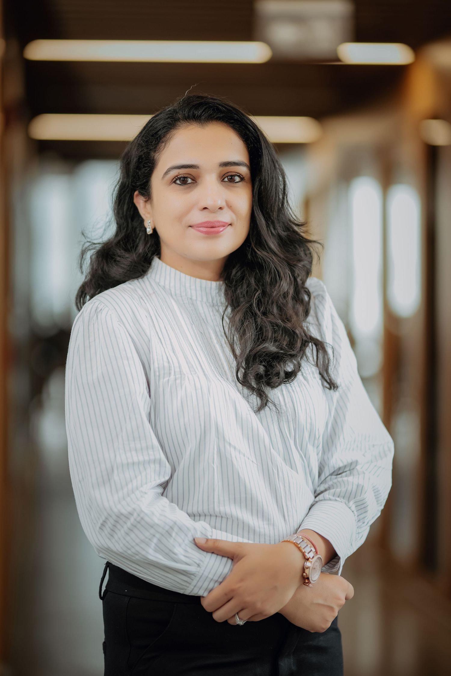 Professional woman in striped white blouse posing confidently in modern office corridor with bokeh background.