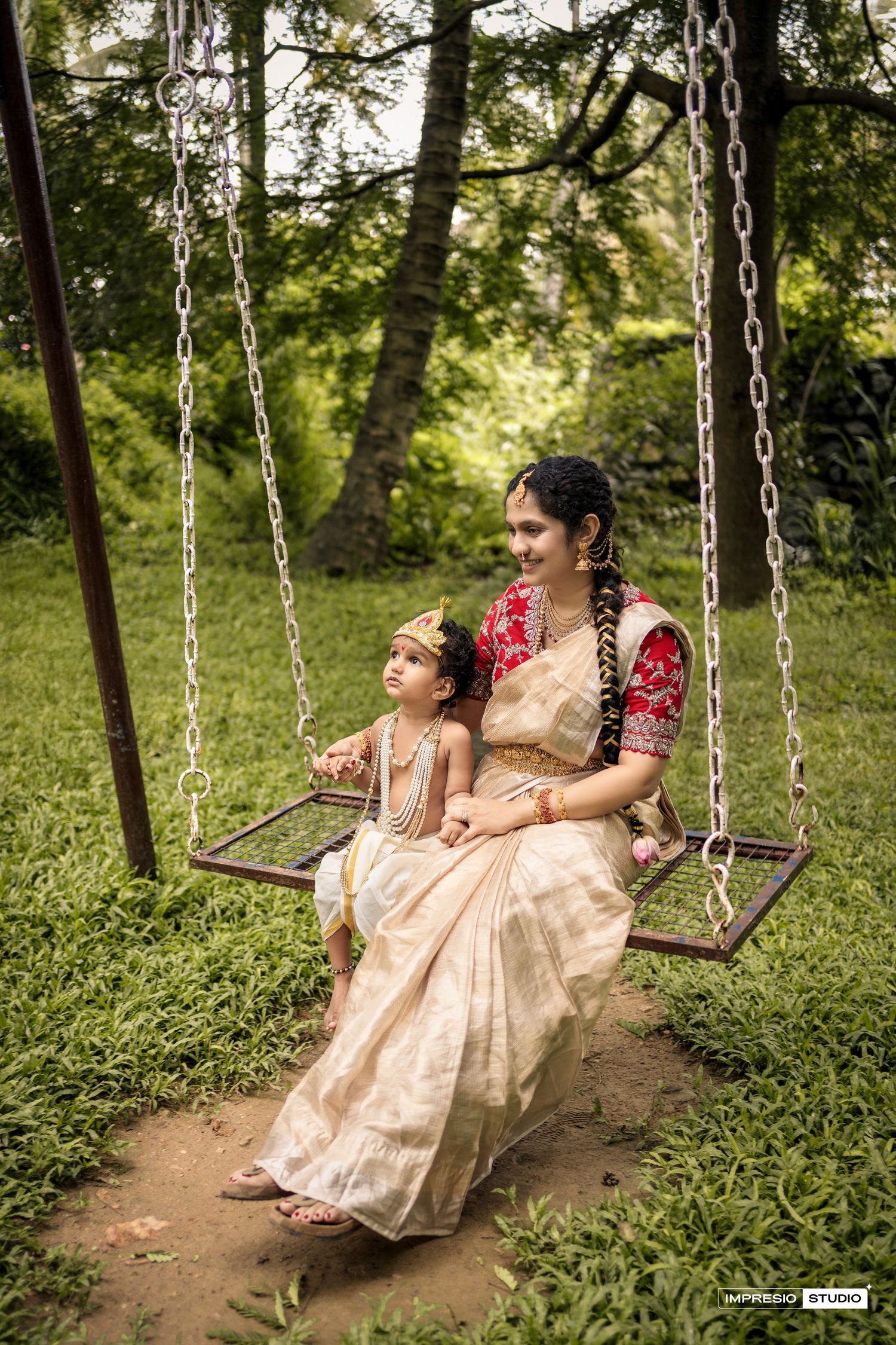 Two people in traditional Indian attire sit together on a wooden swing in a lush green garden setting.