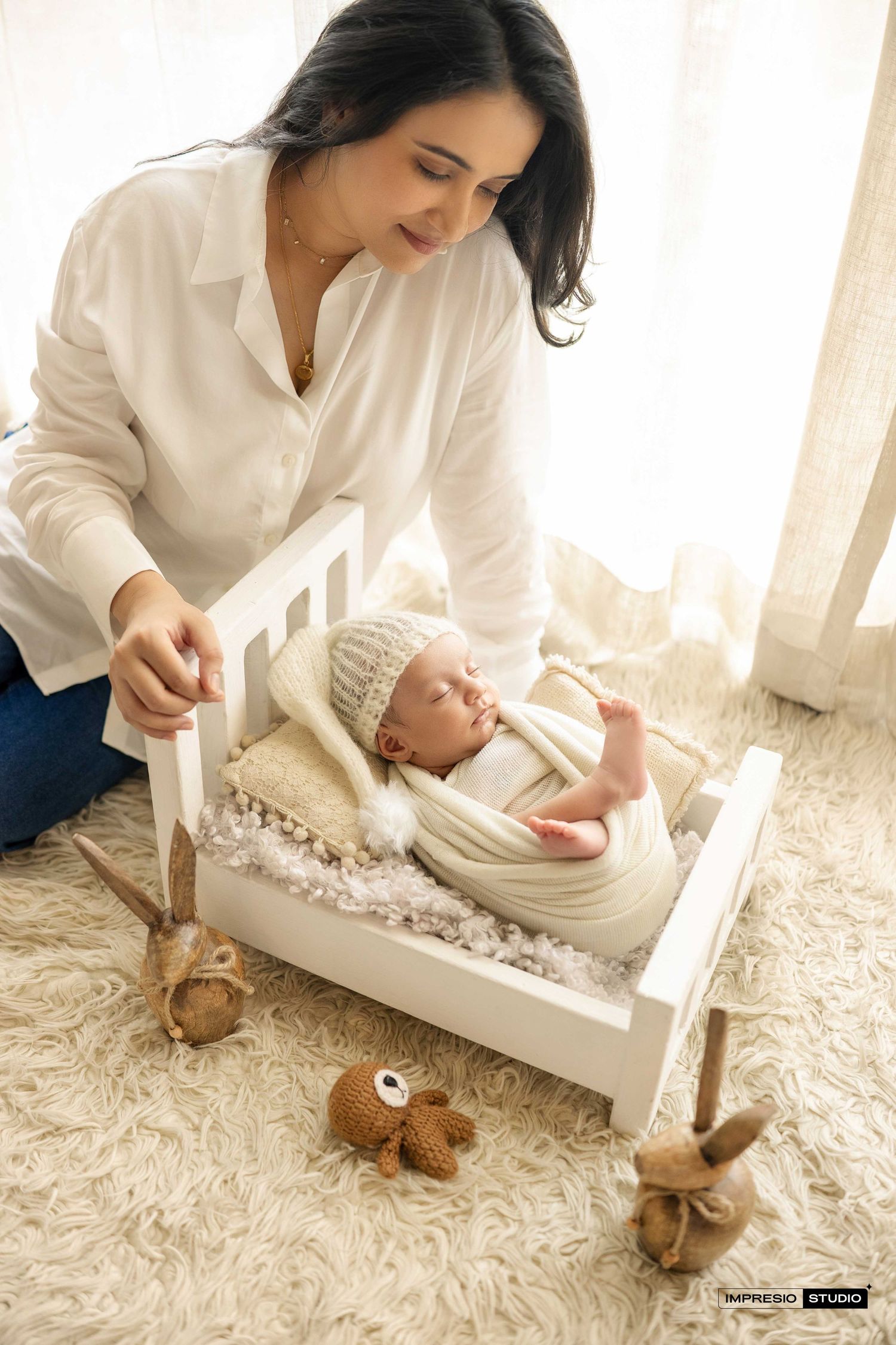 A mother in a white shirt lovingly gazes at her newborn in a tiny white crib, surrounded by plush toys on a fluffy rug.