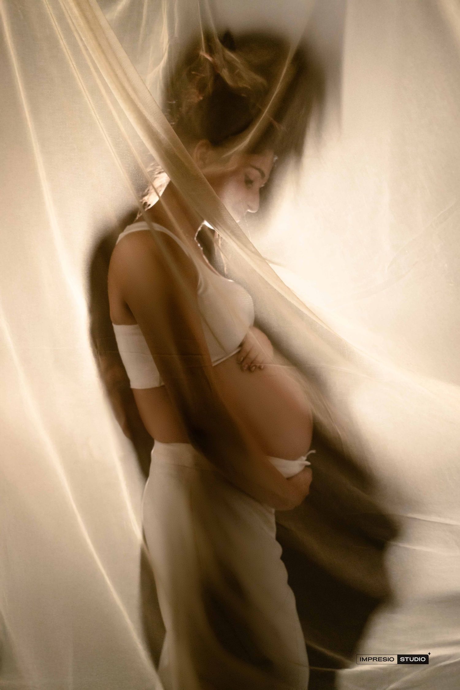 Pregnant woman in white lingerie standing behind sheer flowing curtains in a soft, artistic maternity photo.