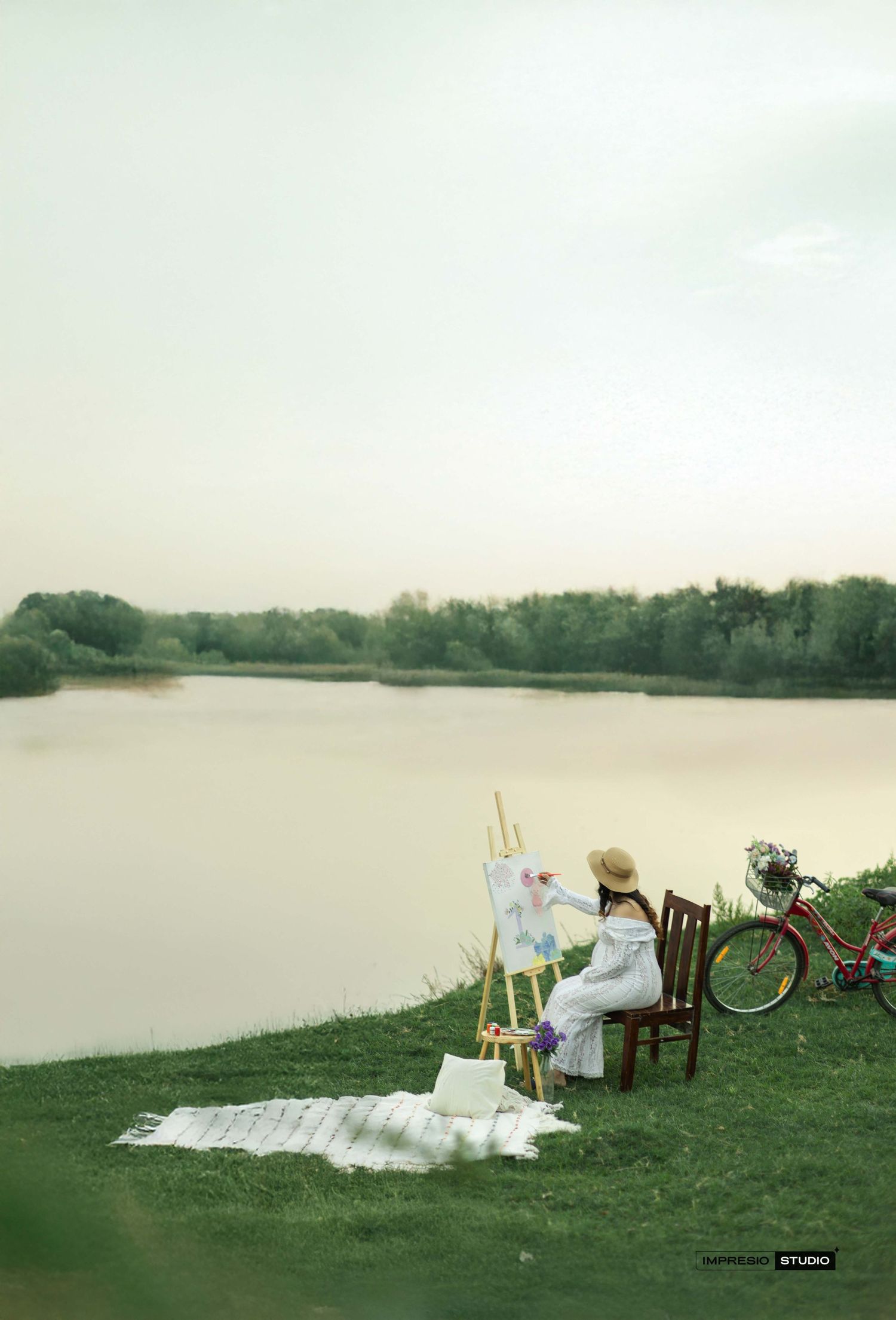 A woman in a white dress and hat paints on an easel by a serene lake, with a red bicycle nearby on lush green grass.