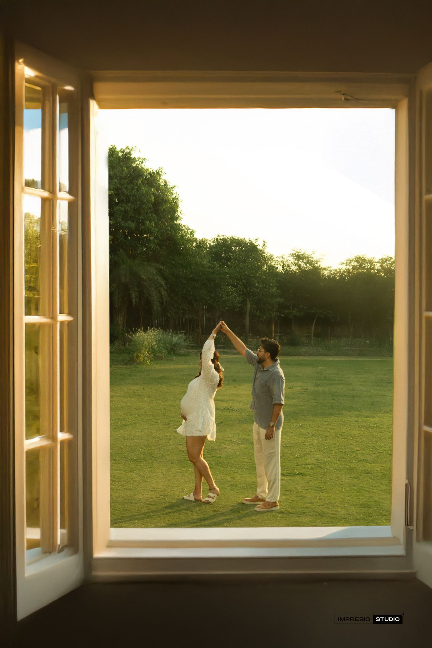 Couple dancing outdoors on green lawn at golden hour, viewed through open white French window frame.