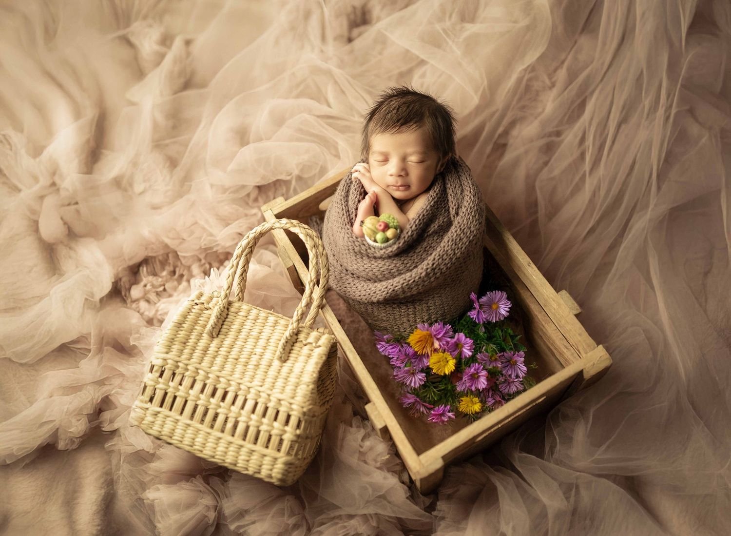 Sleeping newborn baby wrapped in brown knit swaddle inside wooden tray with colorful flowers beside woven basket on beige tulle.