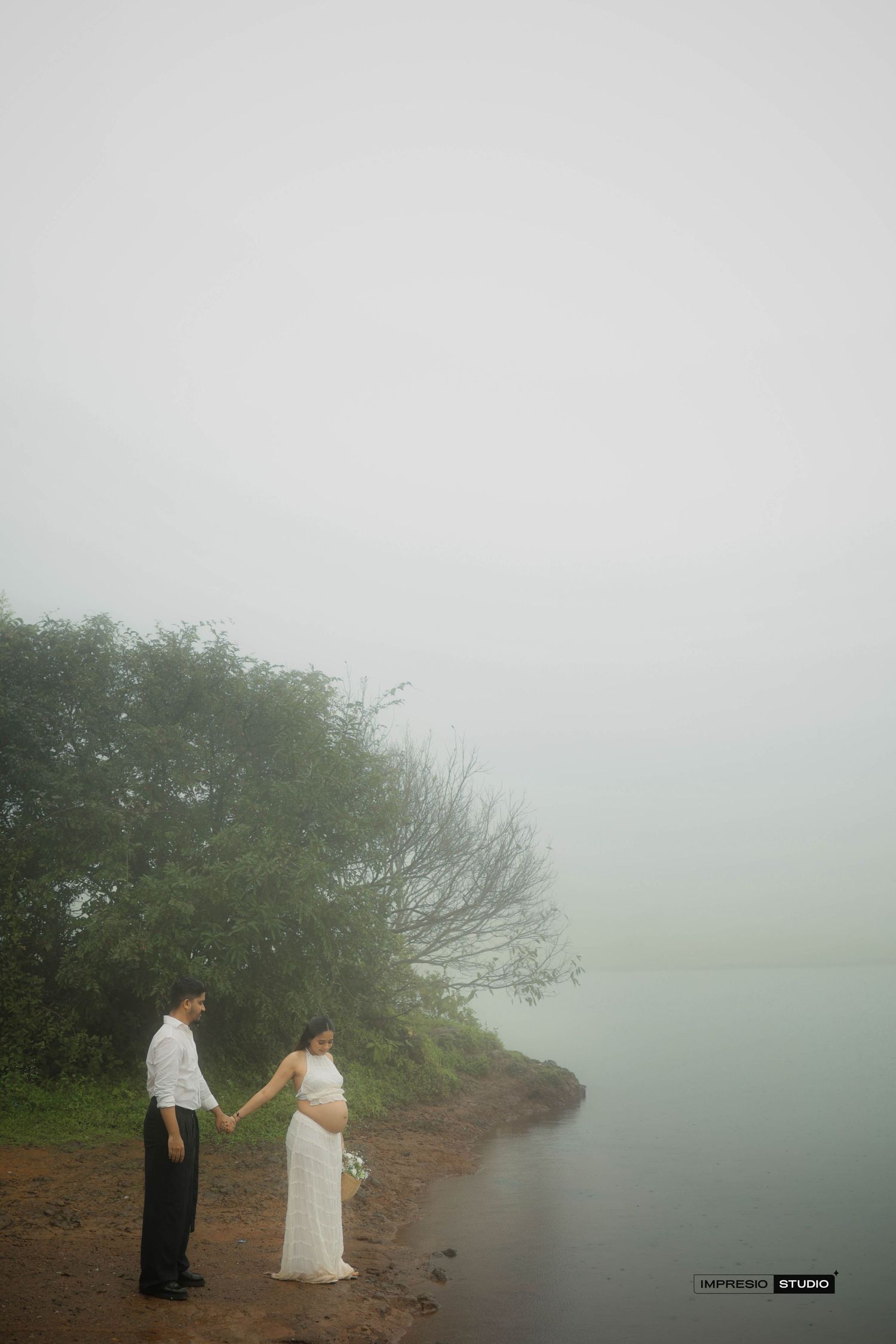 Couple holding hands by a misty lakeside, pregnant woman in white dress, foggy romantic maternity photo shoot outdoors.