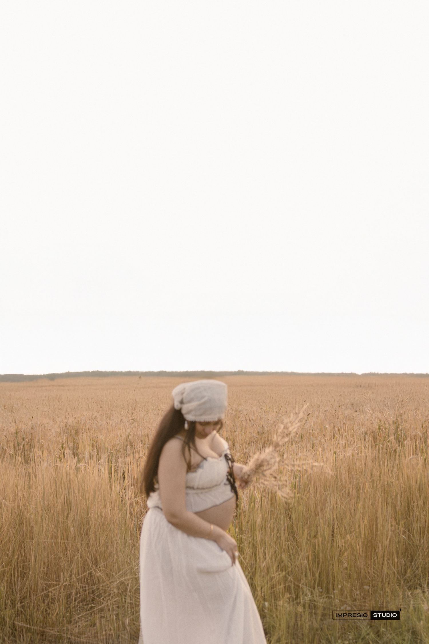 Pregnant woman in white skirt and beanie holding wheat stalks in a golden field during a maternity photo shoot.