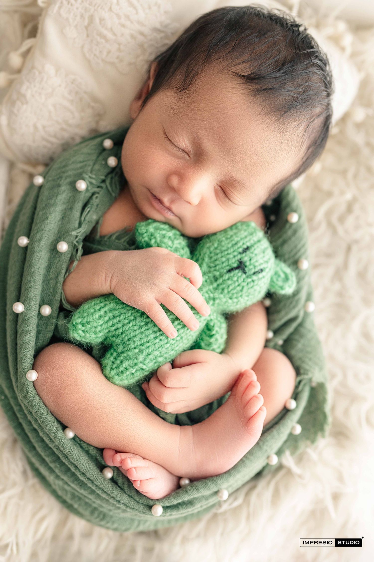 Sleeping newborn baby wrapped in green cloth with pearl details, cuddling a green knitted teddy bear.