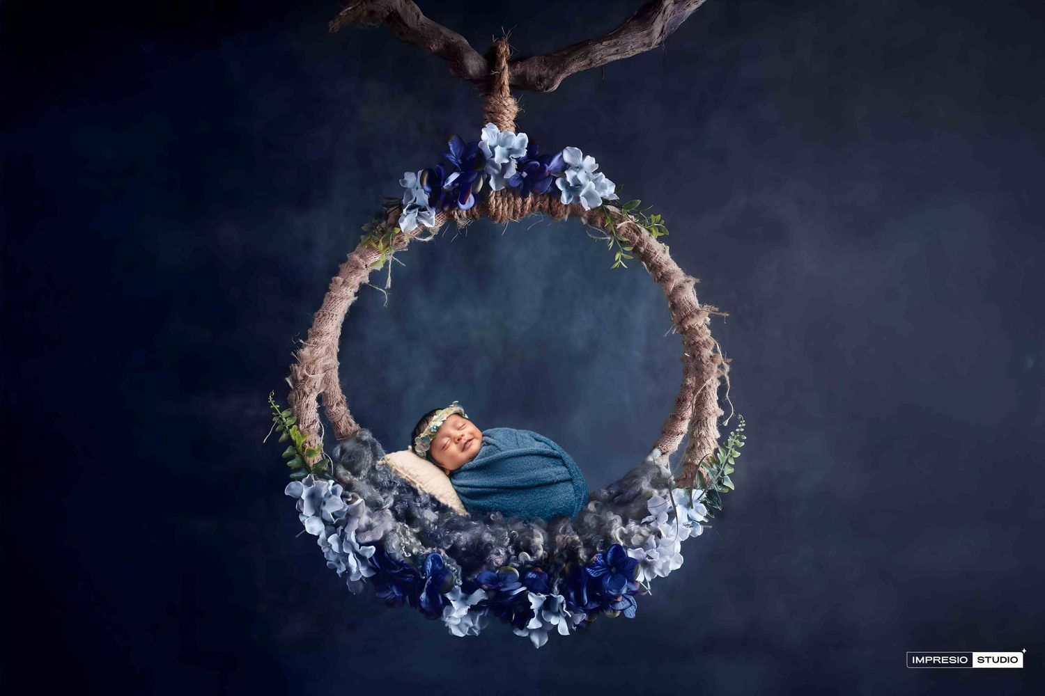Newborn baby sleeping peacefully in a floral wreath swing adorned with blue flowers, suspended against a dreamy dark background.