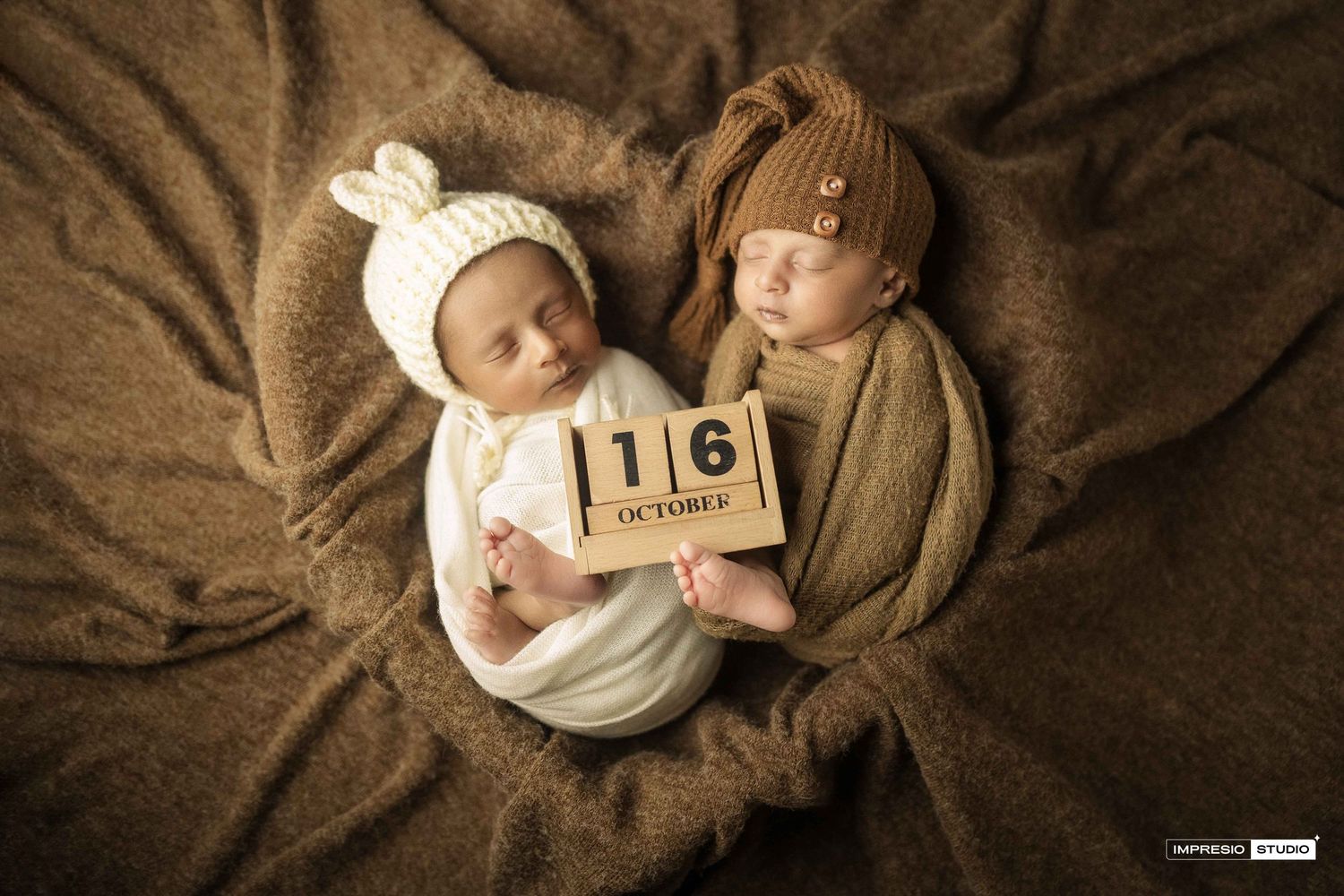Two newborn twins in knit hats lying on brown blanket holding wooden blocks showing October 16 birth date.