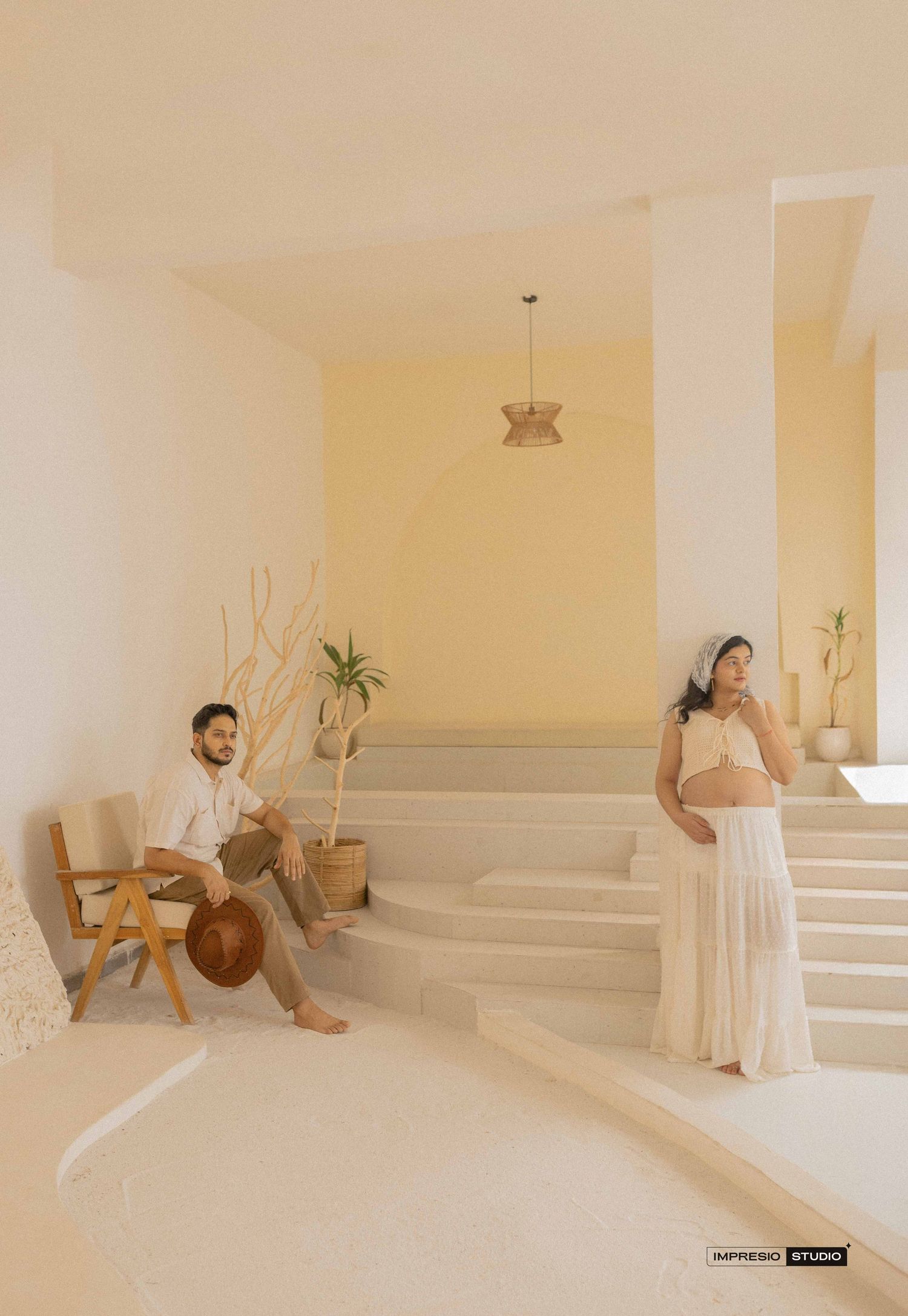 A couple poses in a minimalist beige space, man seated on steps, pregnant woman standing in flowing white skirt.