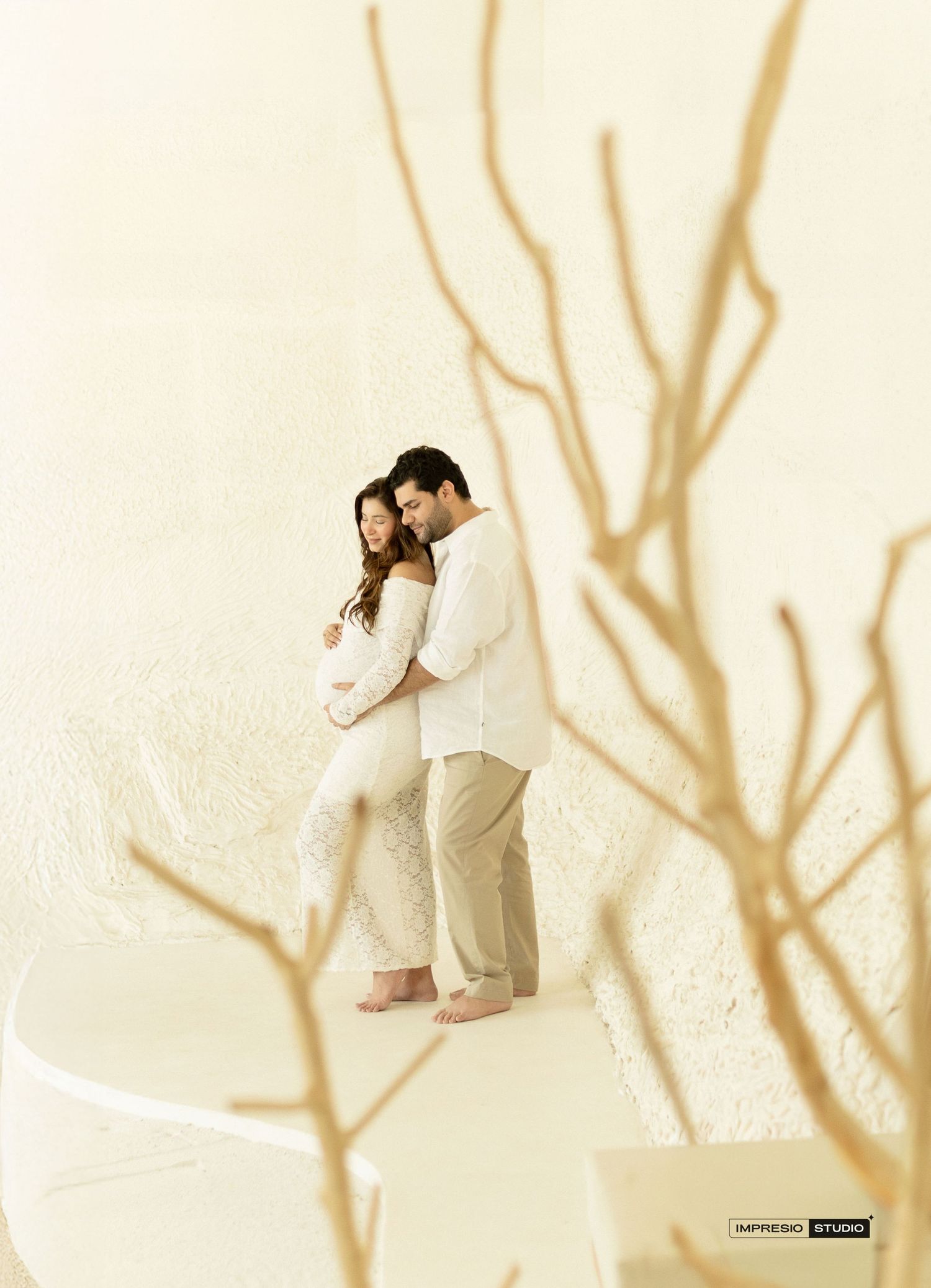 Couple in white outfits embracing tenderly in a minimalist studio with bare branches, maternity photography session.