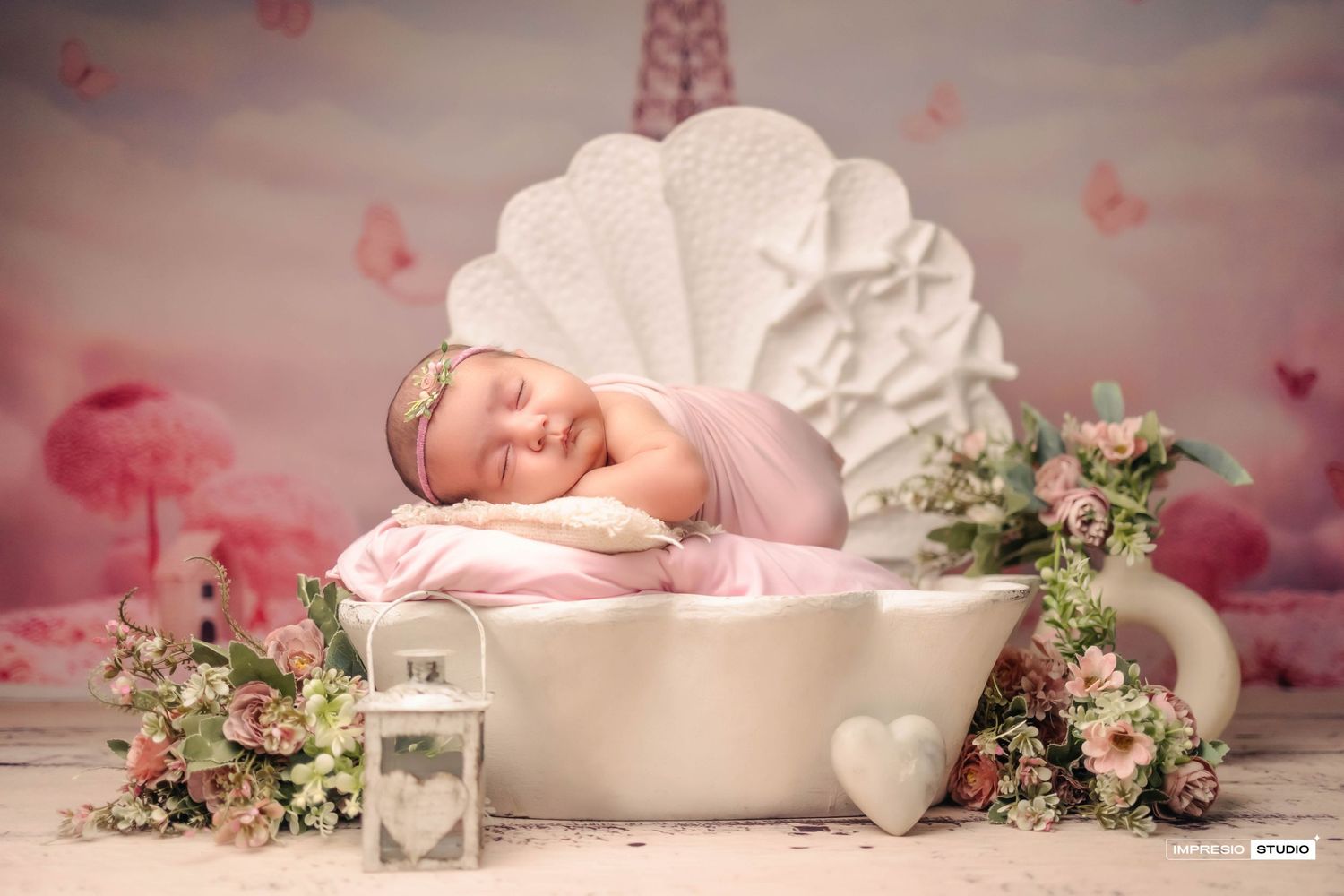 Newborn baby sleeping peacefully in a white teacup prop surrounded by flowers and a pink fairy tale backdrop.
