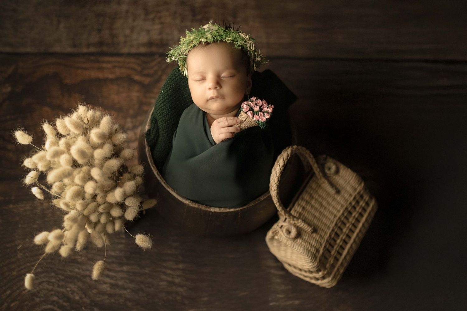 Newborn baby sleeping in a green bucket with a floral crown, holding pink flowers, beside dried botanicals and a wicker purse.
