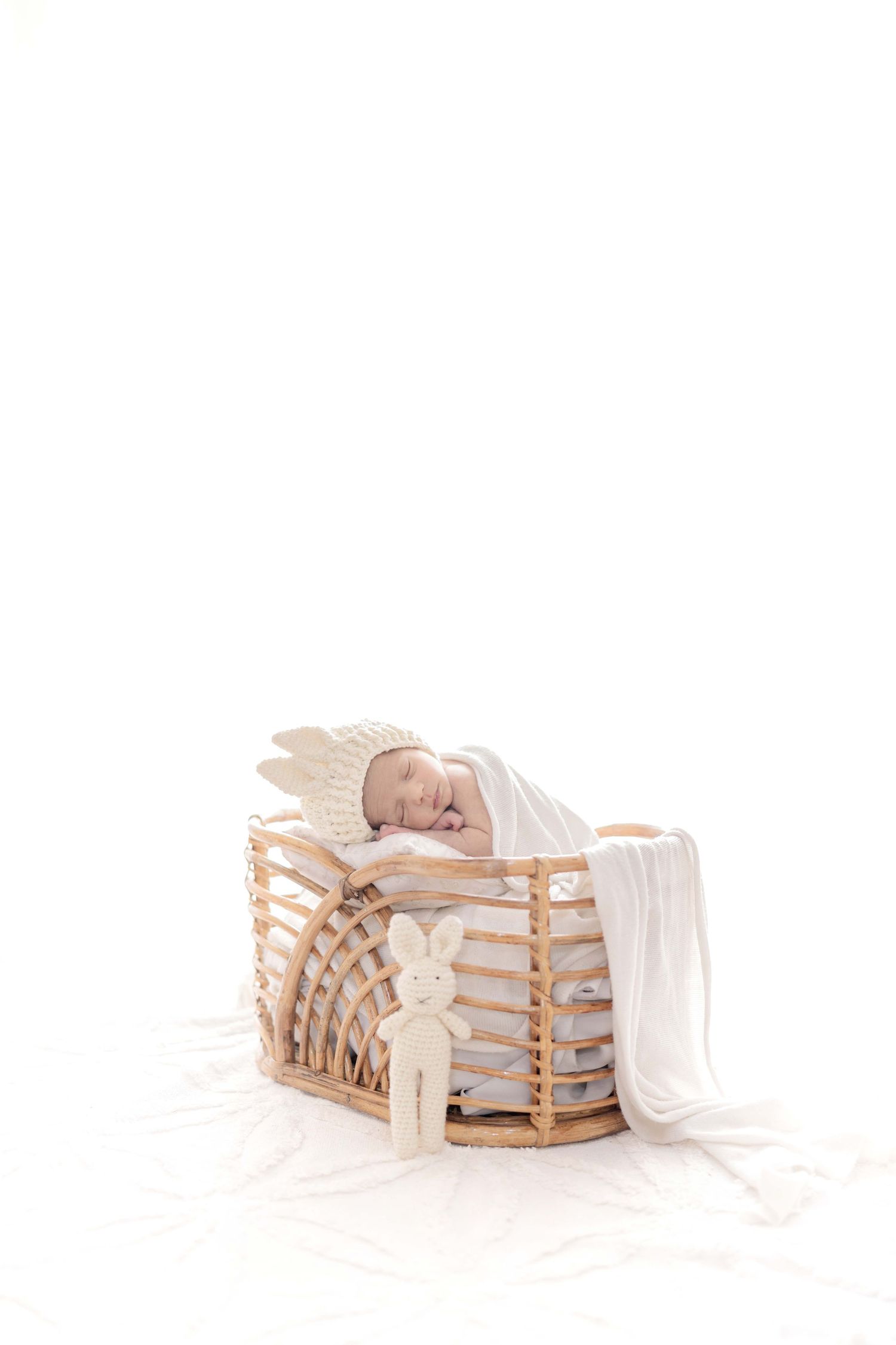 Newborn baby sleeping peacefully in a wicker bassinet with a white blanket and stuffed bunny toy beside it.