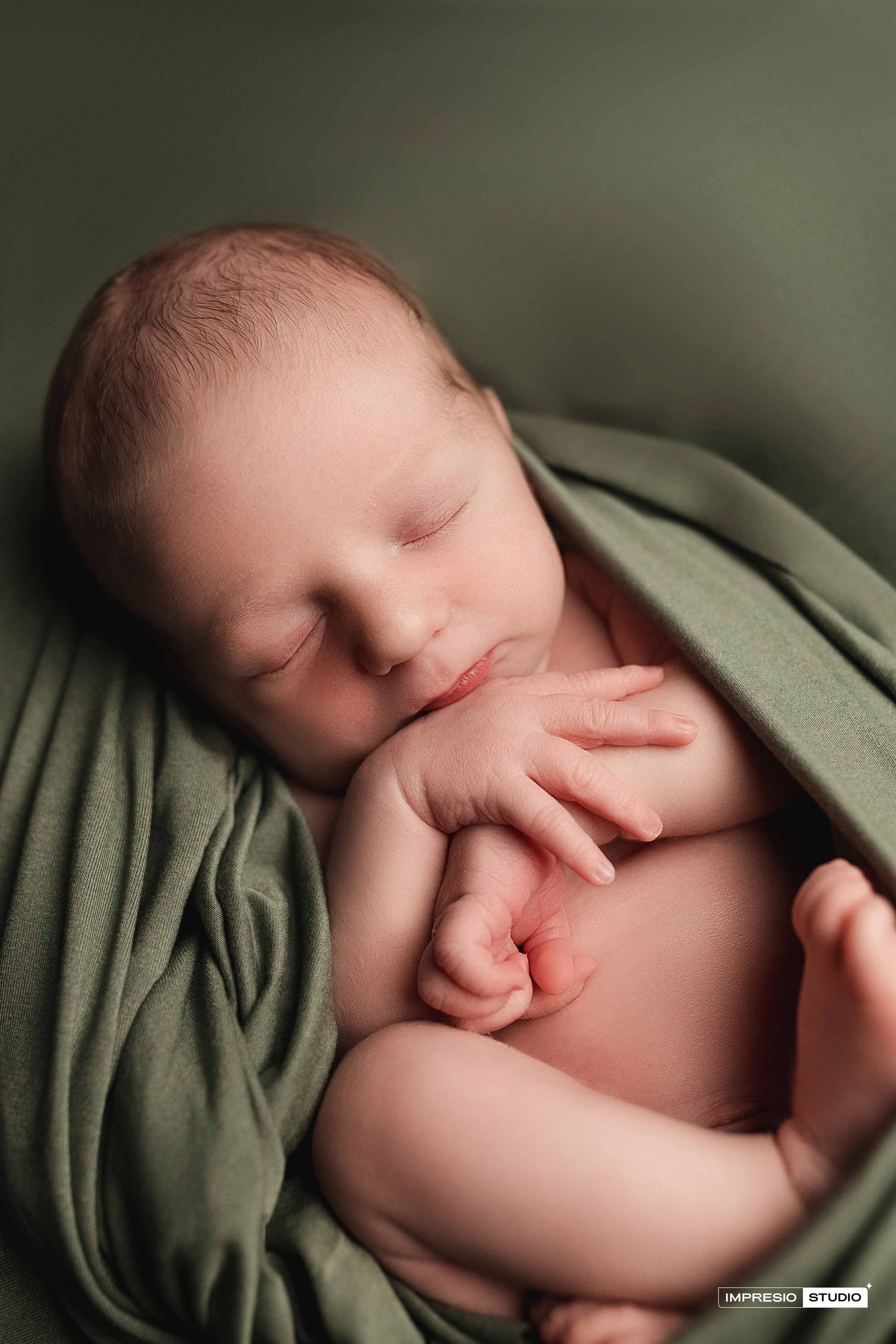 Sleeping newborn baby wrapped in soft green fabric, peacefully curled with tiny hands visible.