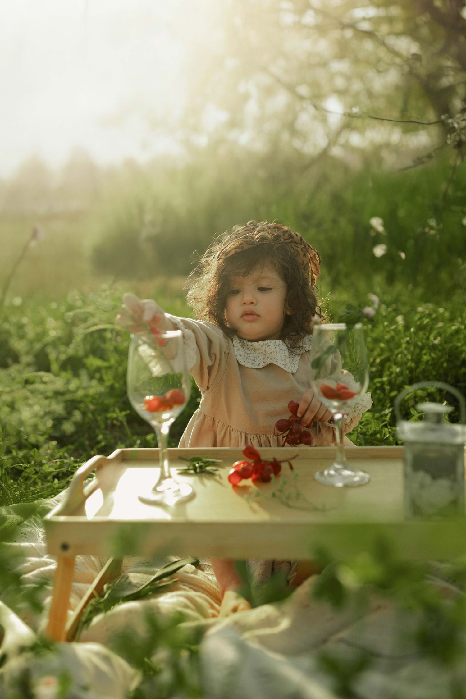 Young girl in pink dress playing with wine glasses and red flowers on a wooden tray outdoors in sunlit garden.