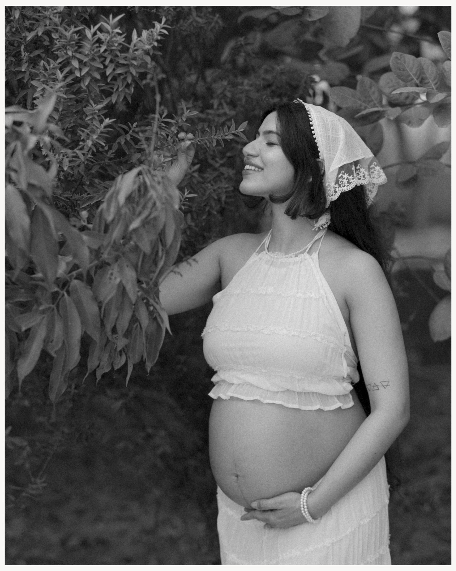 Black and white maternity photo of a smiling pregnant woman in white dress holding her belly near lush green foliage.