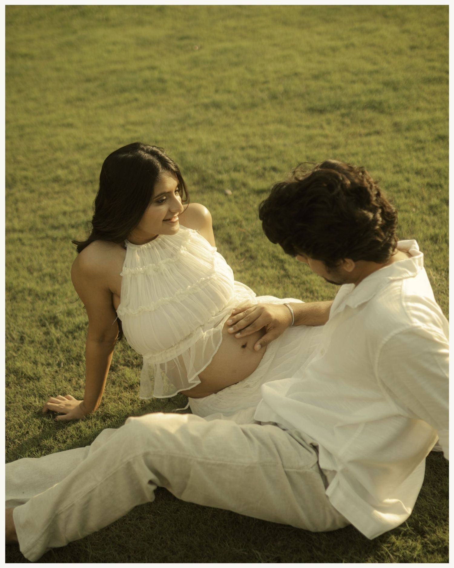 A pregnant woman in white dress sits on grass with partner touching her baby bump during maternity photoshoot.