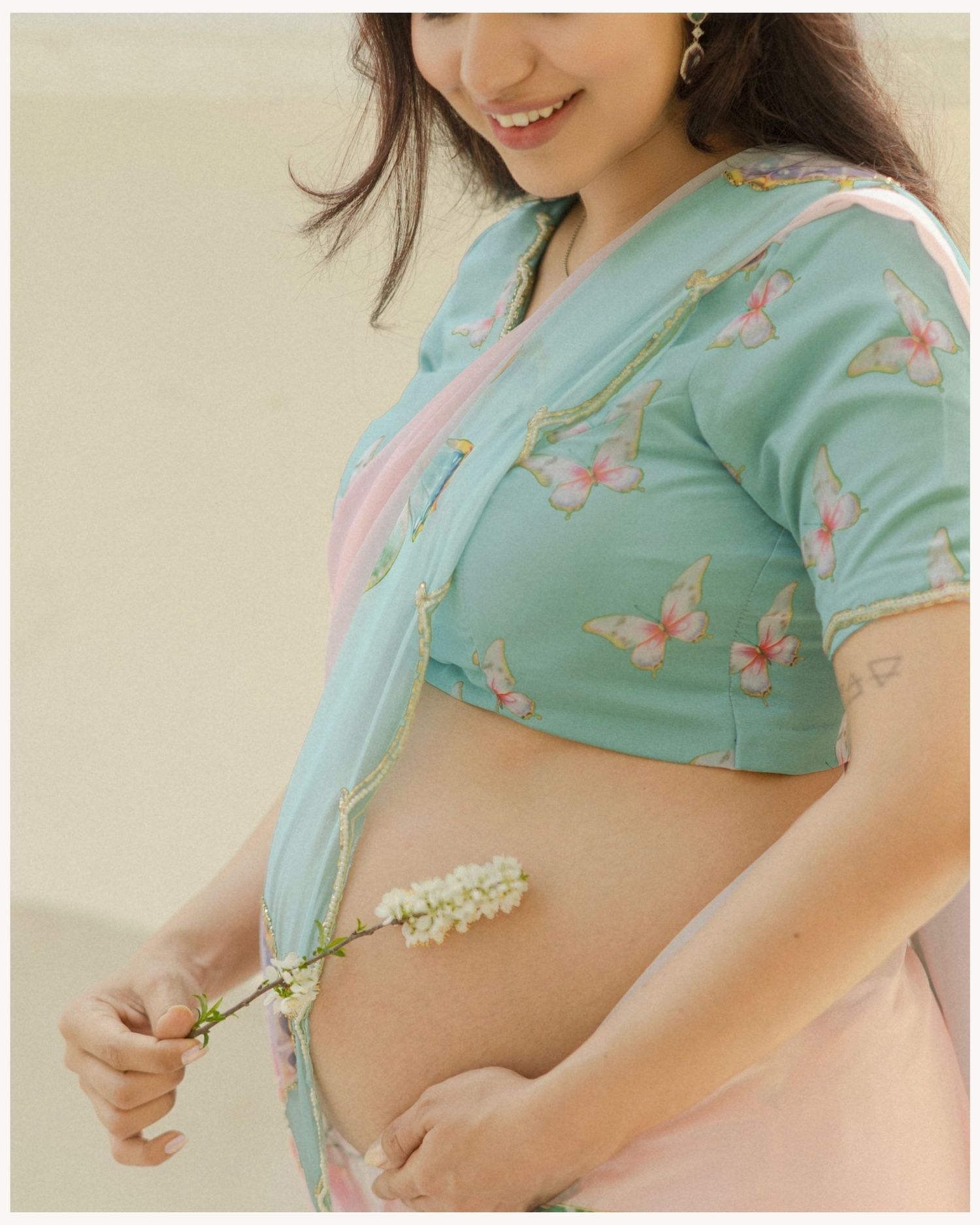 Smiling pregnant woman in teal butterfly print crop blouse and pink saree holding white flowers against her baby bump.