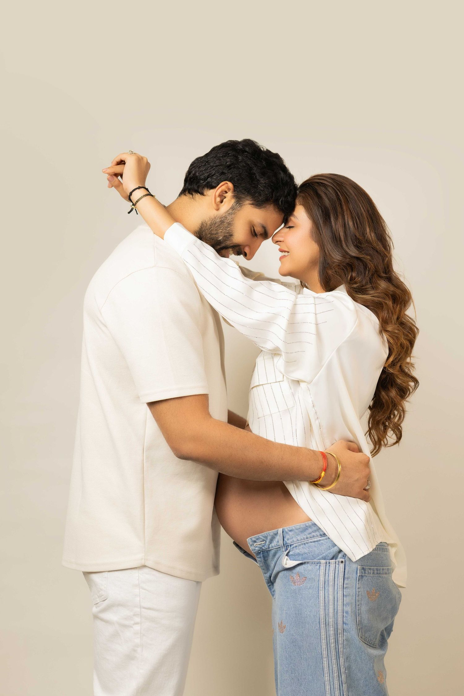 Couple embracing tenderly, pregnant woman in jeans and white top with partner in white outfit on neutral background.