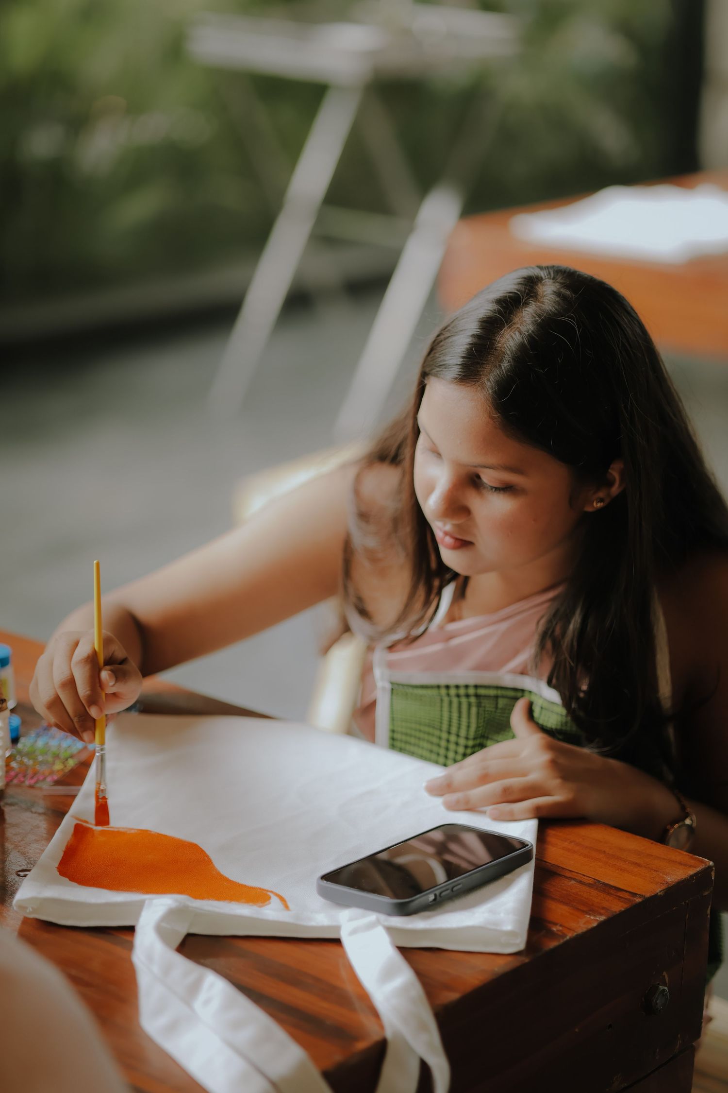 Young girl painting on a white tote bag with an orange paintbrush at a wooden table.