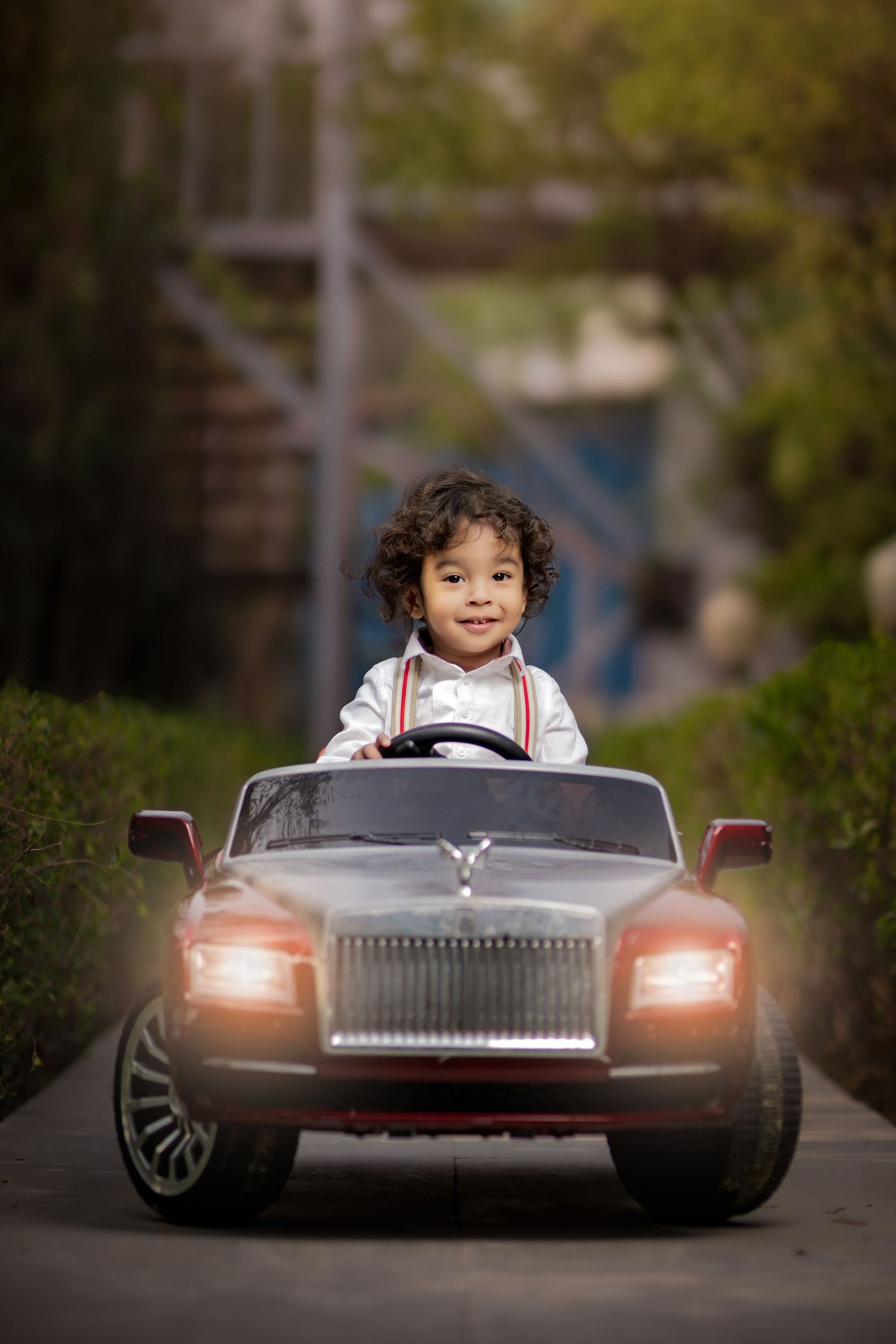 Young child with curly hair smiling while driving a toy Rolls-Royce electric car outdoors.