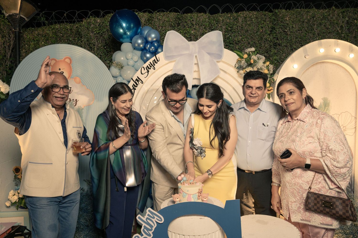 Group of five people cutting a cake at a blue-themed birthday celebration with balloon decorations.
