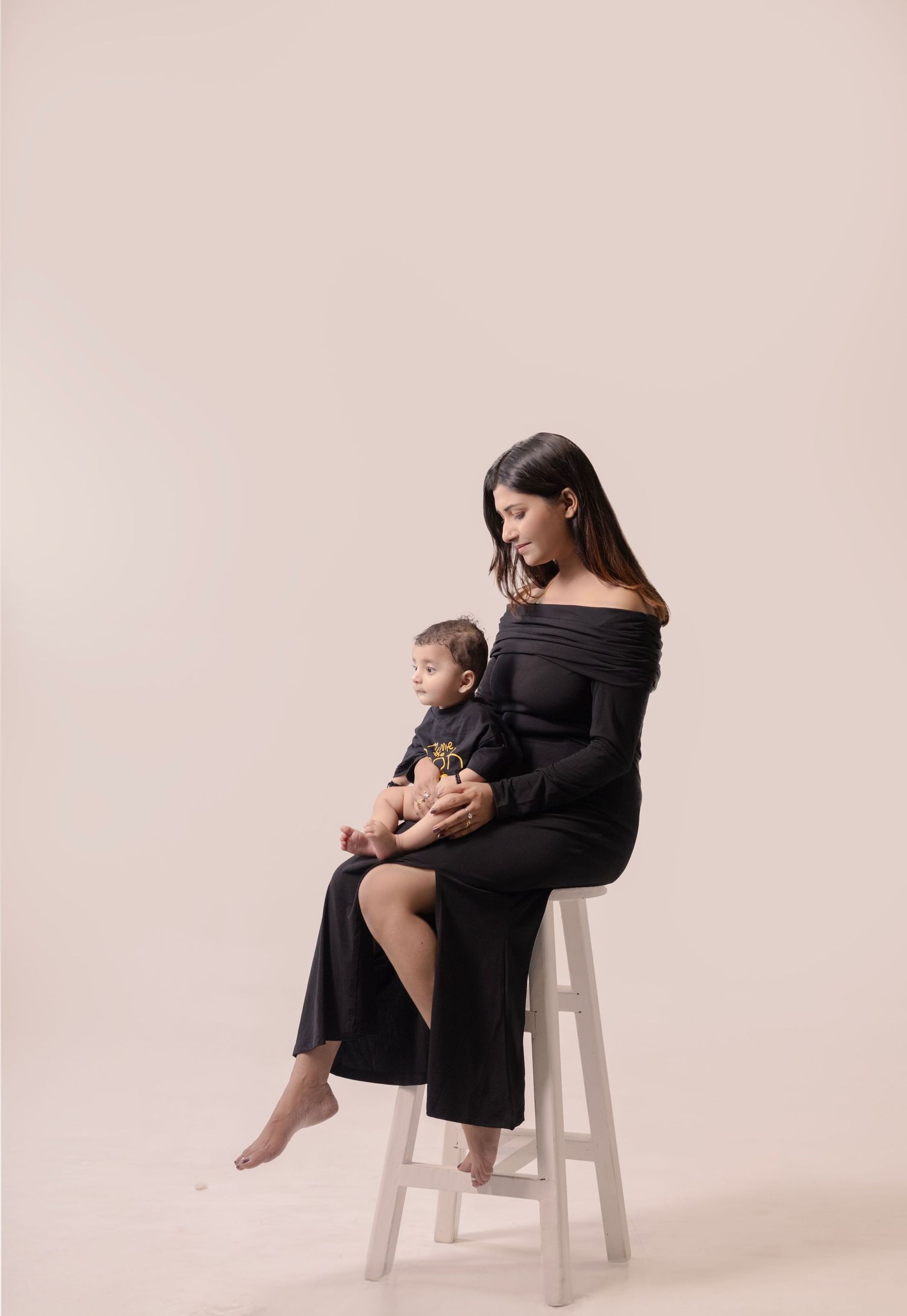 A woman in a black off-shoulder jumpsuit sits on a white stool holding a baby dressed in matching black outfit.