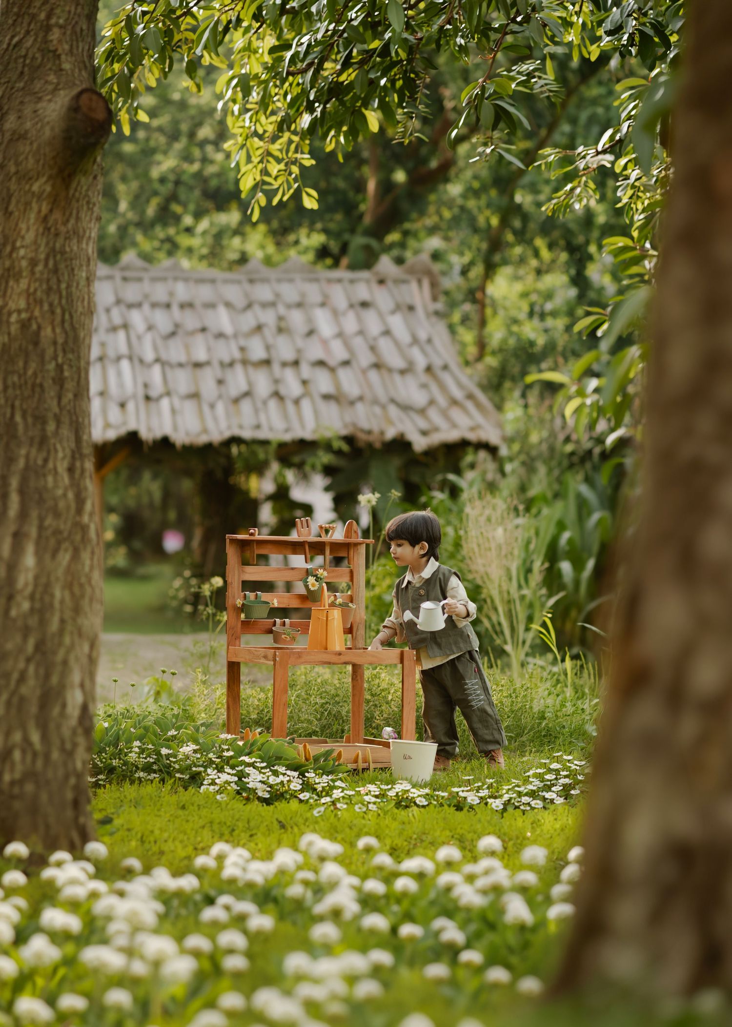 Young child playing outdoors near a wooden shelf and bucket, surrounded by white flowers and lush green trees.