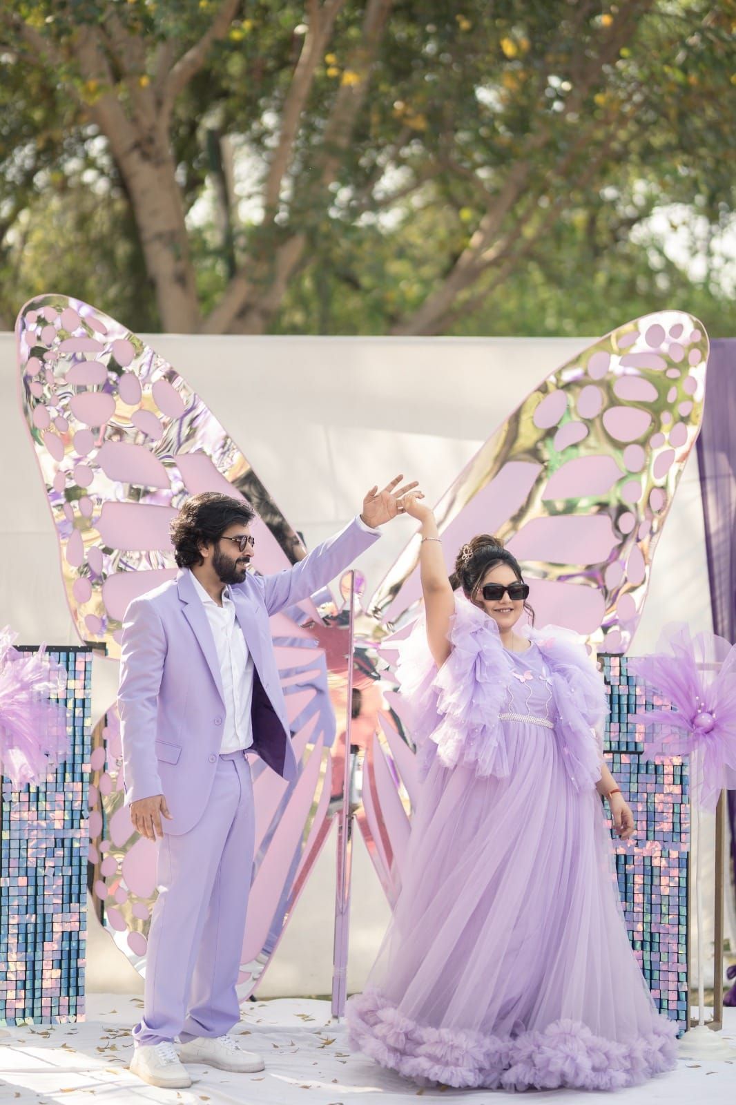 Couple in matching lavender outfits posing in front of a large decorative butterfly backdrop at an outdoor event.