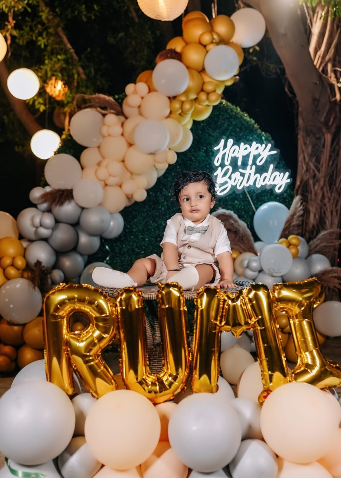 Baby boy sits among gold letter balloons spelling RUMO, surrounded by white and gray balloon garland with Happy Birthday neon sign.