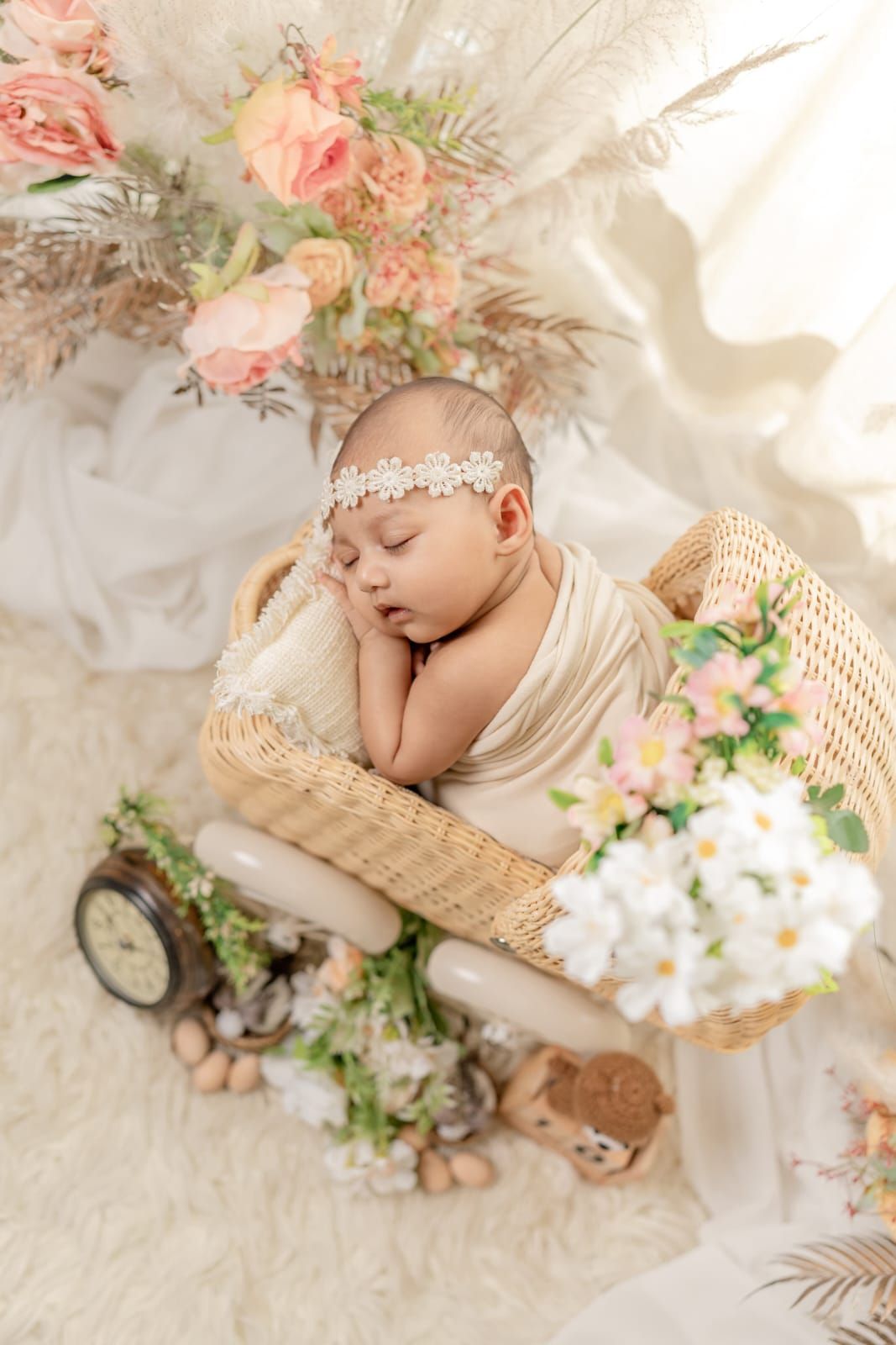 Sleeping newborn baby wrapped in cream swaddle with floral headband resting in wicker basket surrounded by flowers.