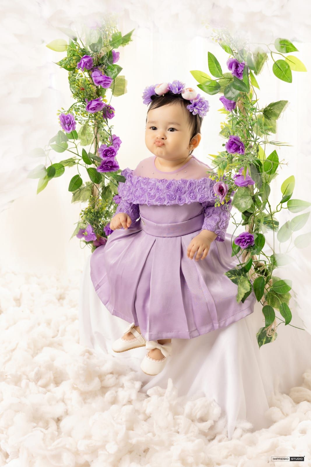 Baby girl in purple dress and floral crown sits on flower-adorned swing surrounded by white petals.