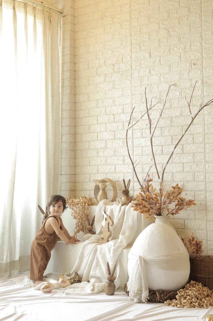 Young child in brown romper plays beside boho-style dried floral decor and white vases in bright studio.