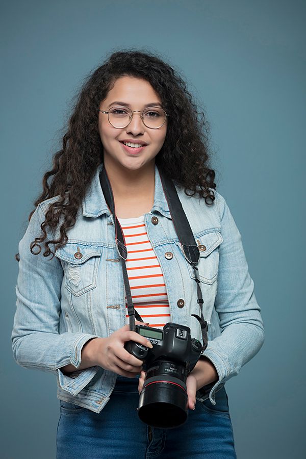 Young woman with curly hair and glasses holding a DSLR camera, wearing a denim jacket over a striped shirt.