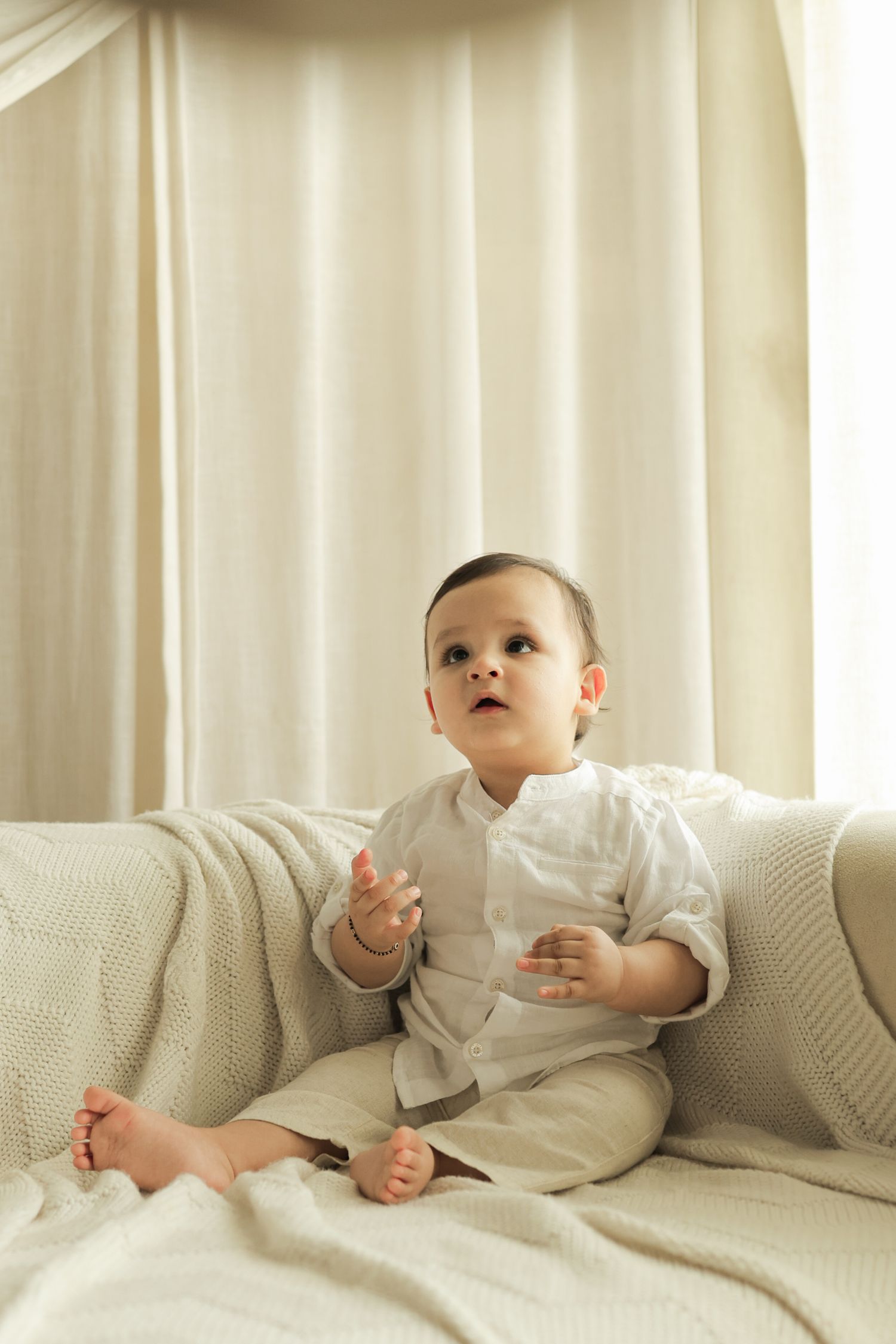 A cute baby boy in a white outfit sits on a cream sofa, looking up curiously in a bright, softly lit room.