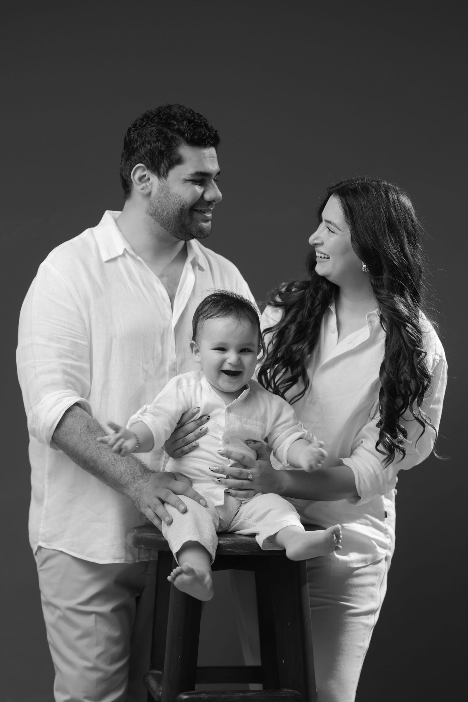 Black and white family portrait of smiling parents in white outfits holding their laughing baby on a stool.