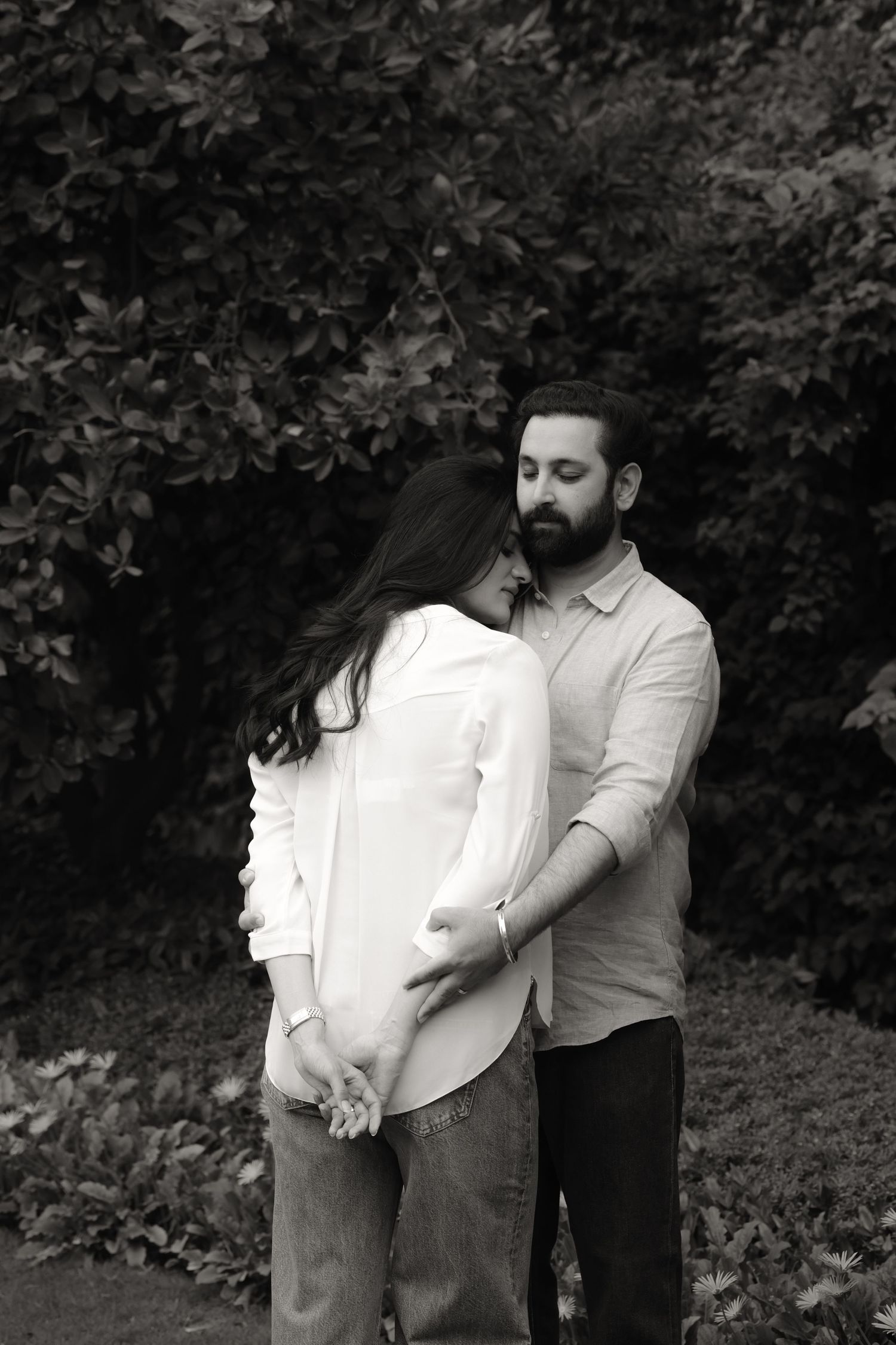 Black and white photo of a couple embracing tenderly outdoors, man resting his head on woman's hair near lush greenery.