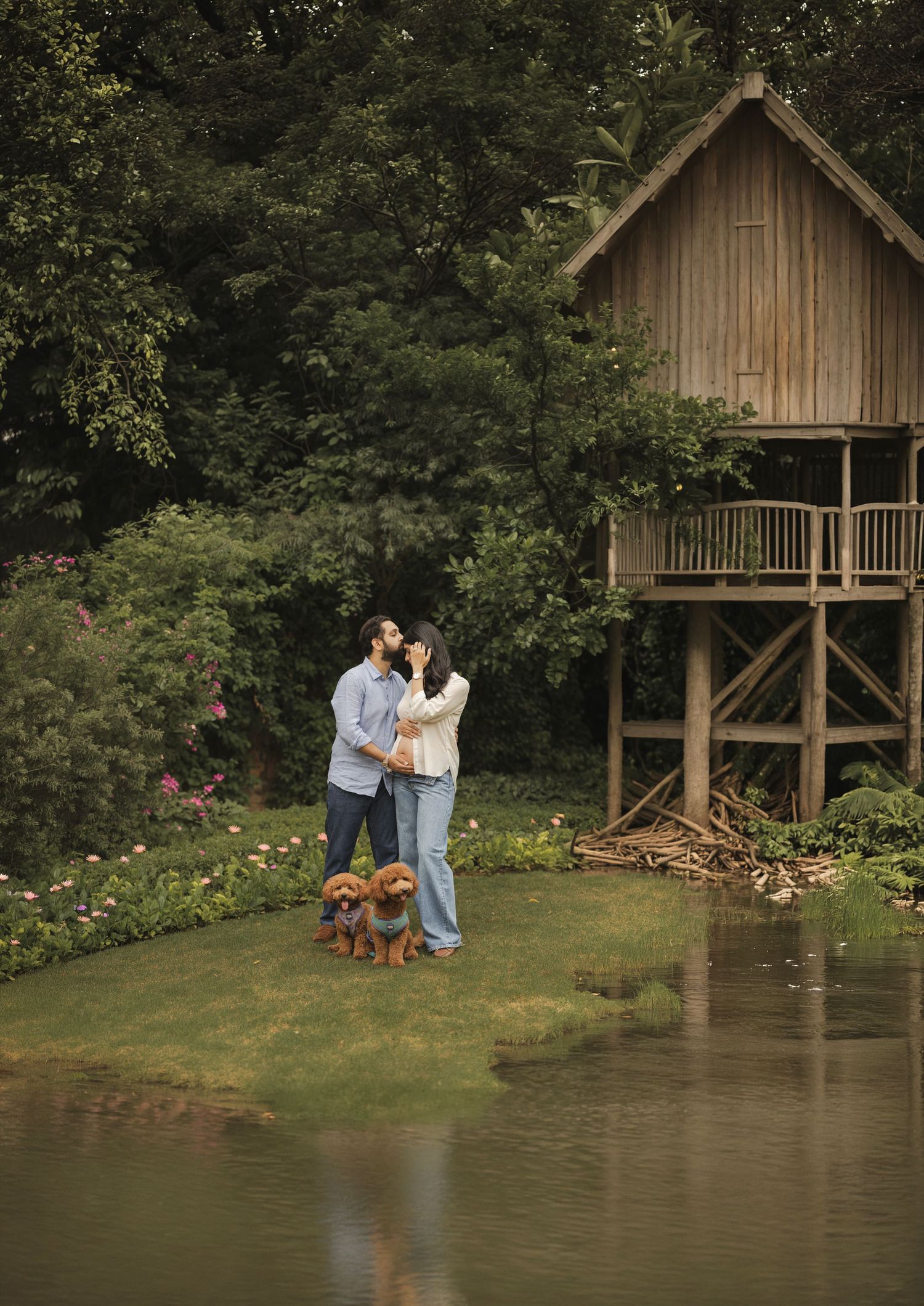 Couple kissing near a rustic treehouse by a pond with two small brown dogs on a lush green property.