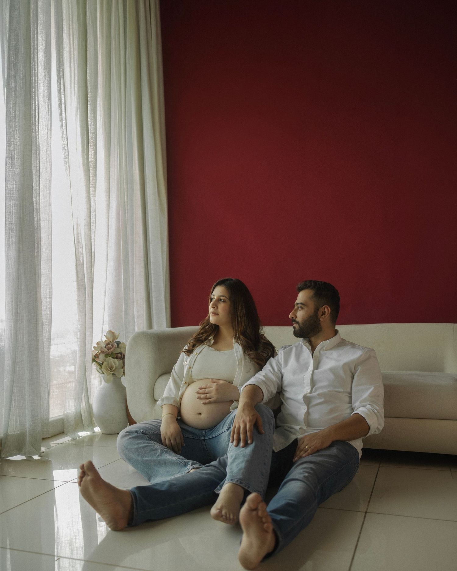 Pregnant couple sitting on floor against red wall, woman in white top holding baby bump, man in white shirt beside her.