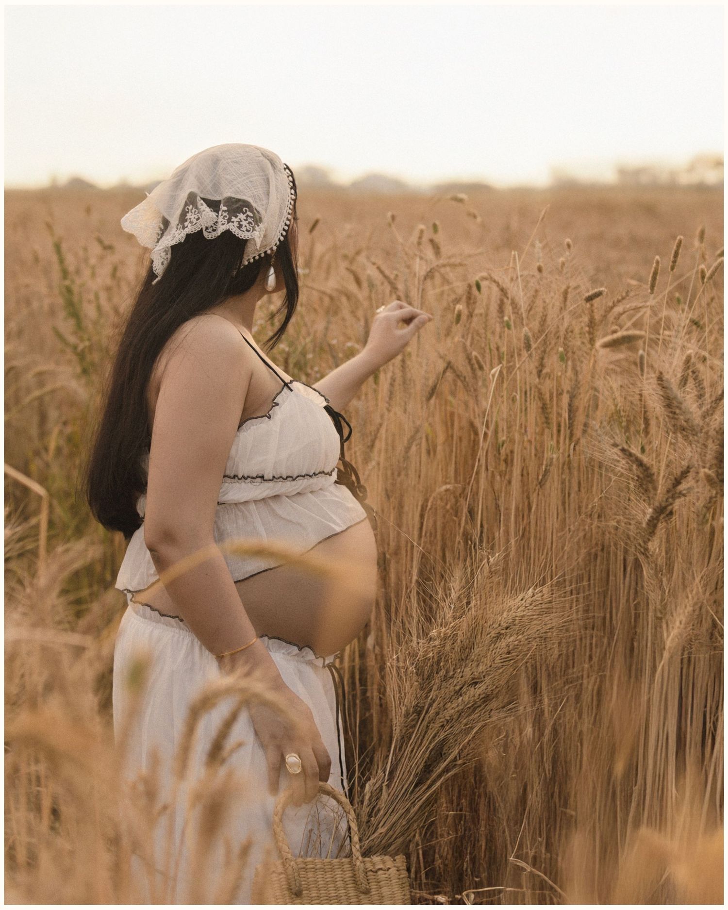 Pregnant woman in white outfit and lace hat touching wheat stalks in a golden field at sunset.