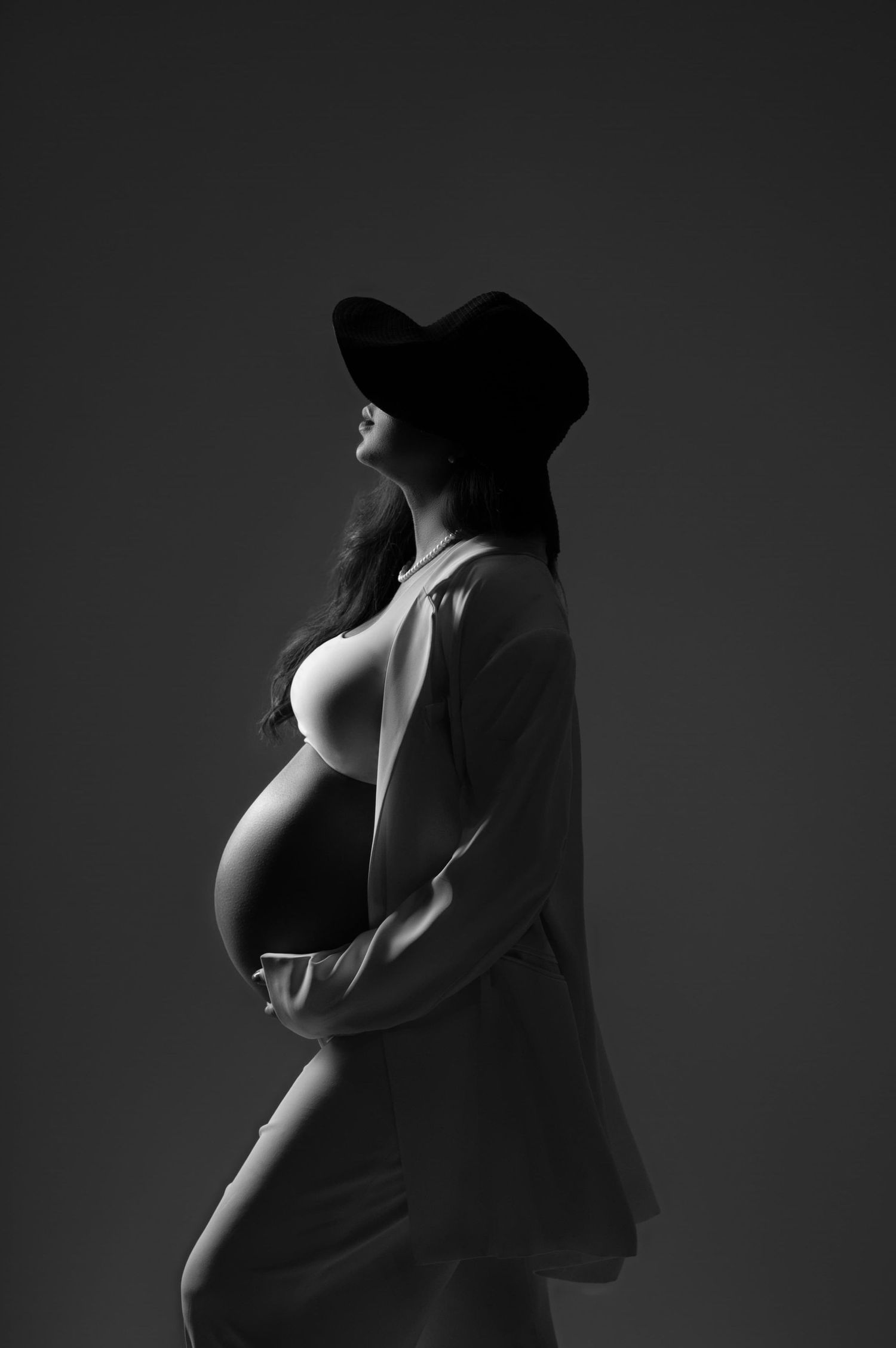 Artistic black and white maternity portrait showing silhouette of pregnant figure in hat against dark background for elegant maternity shoot