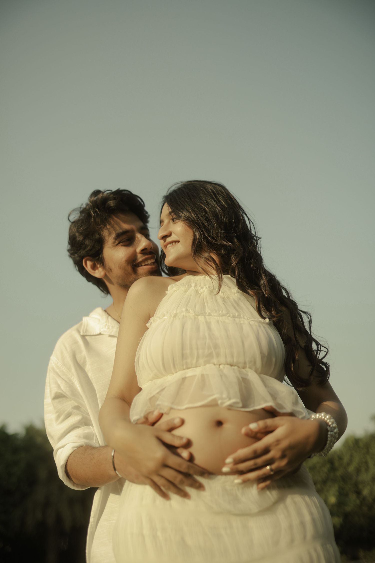 A couple in white clothing shares a tender moment outdoors, with the man embracing the woman's pregnant belly.