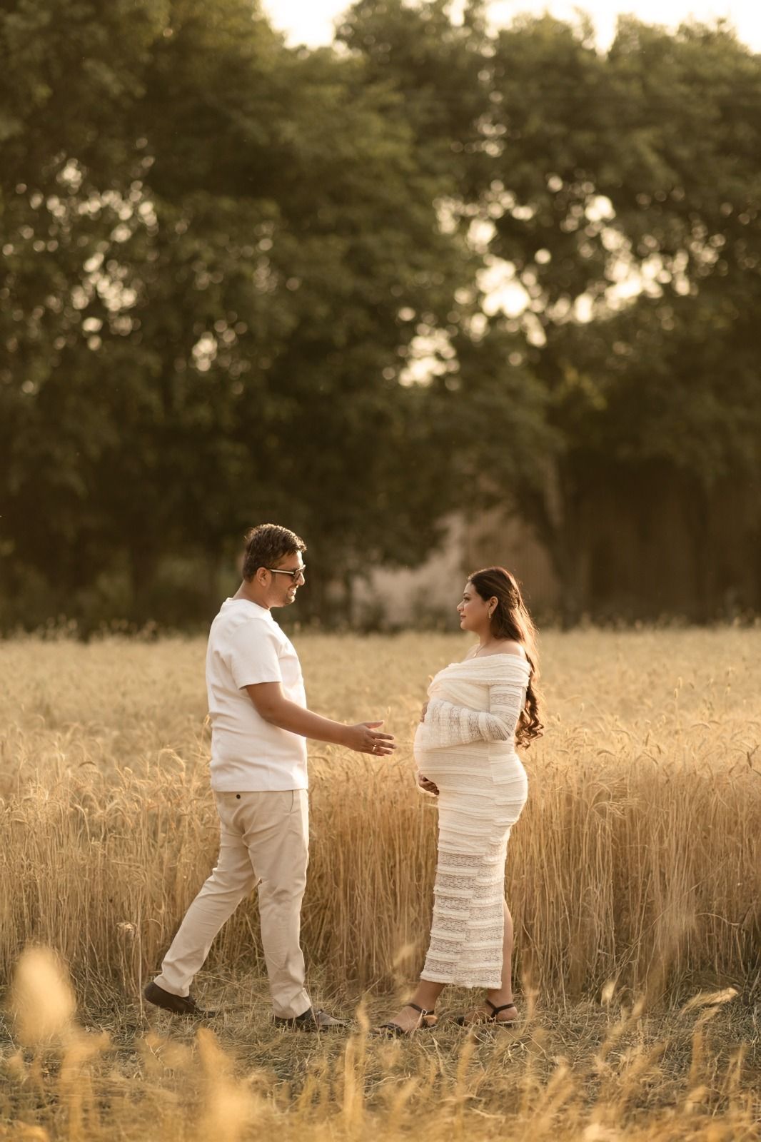 Tender moments between couple walking through sunlit wheat field in white attire.