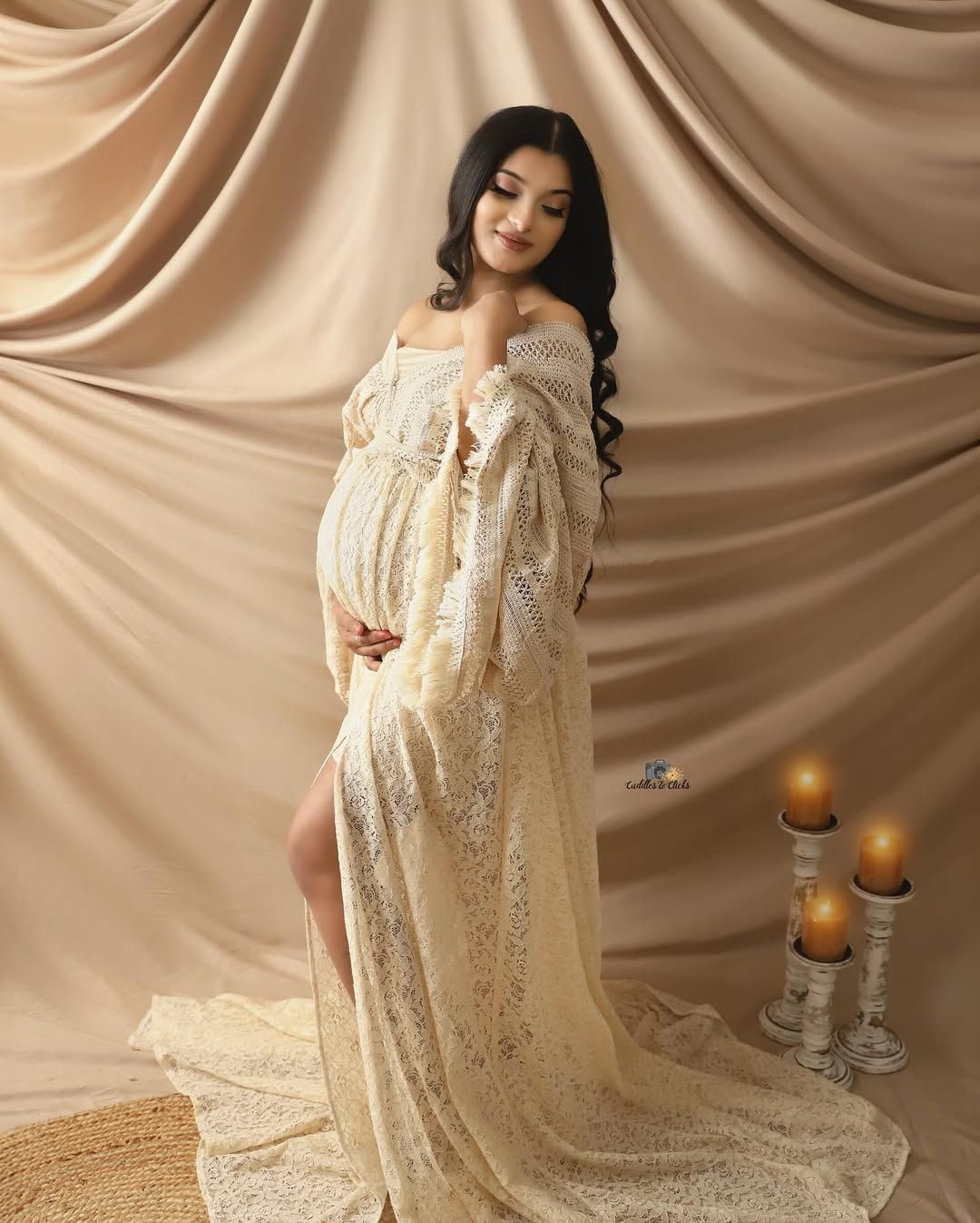 Maternity photo featuring an elegant cream lace gown with off-shoulder design against draped beige backdrop with candles.
