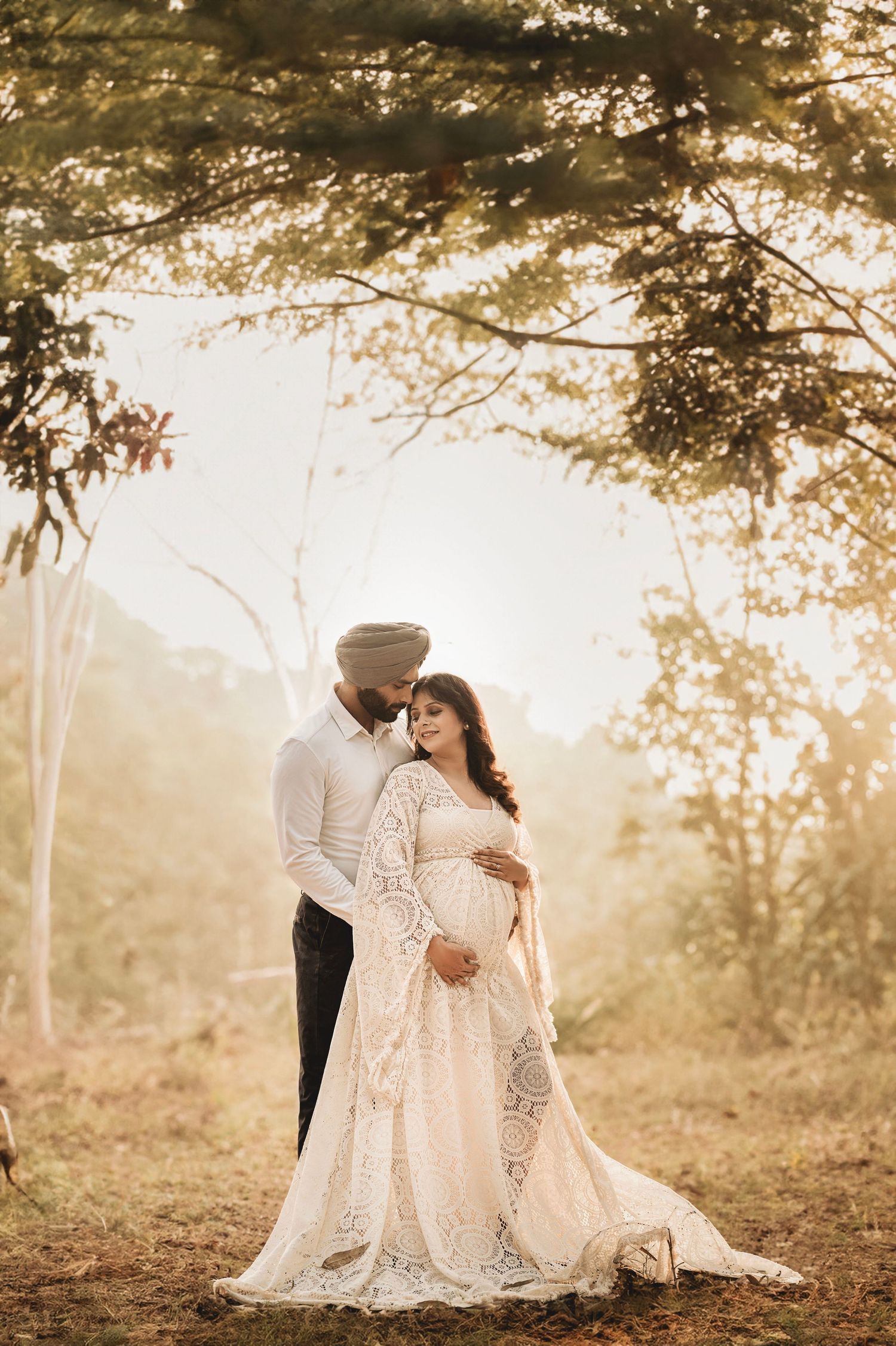 Romantic couple in white attire embraces during a dreamy outdoor photoshoot with golden sunlit trees in background.