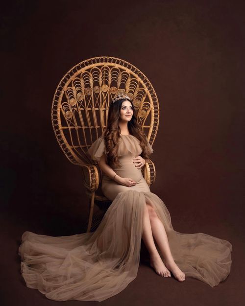 Maternity photo featuring flowing beige tulle gown and ornate peacock chair against dark backdrop.